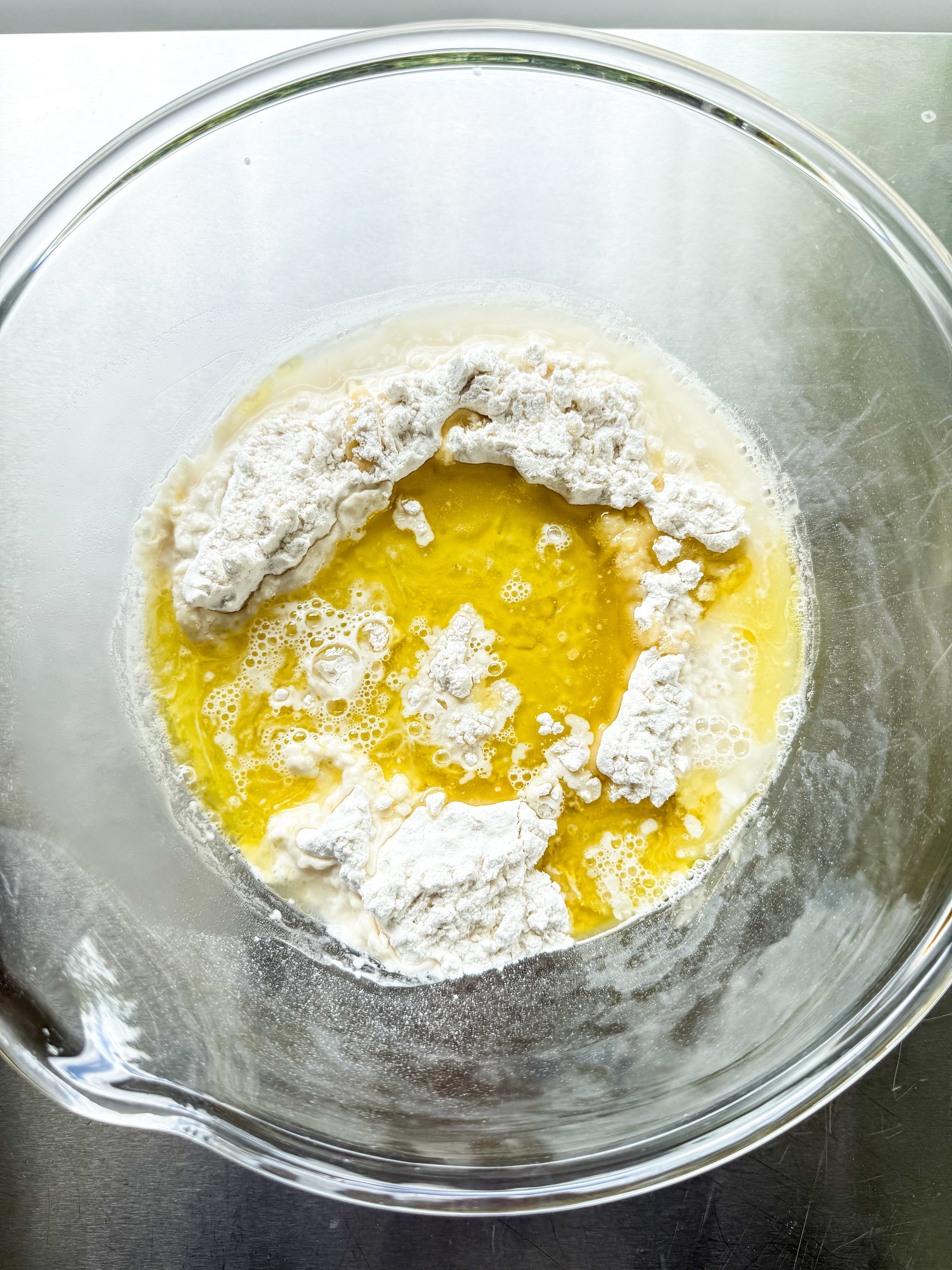 A glass mixing bowl contains flour, melted butter, and milk on a stainless steel surface, ready to be mixed together for baking.