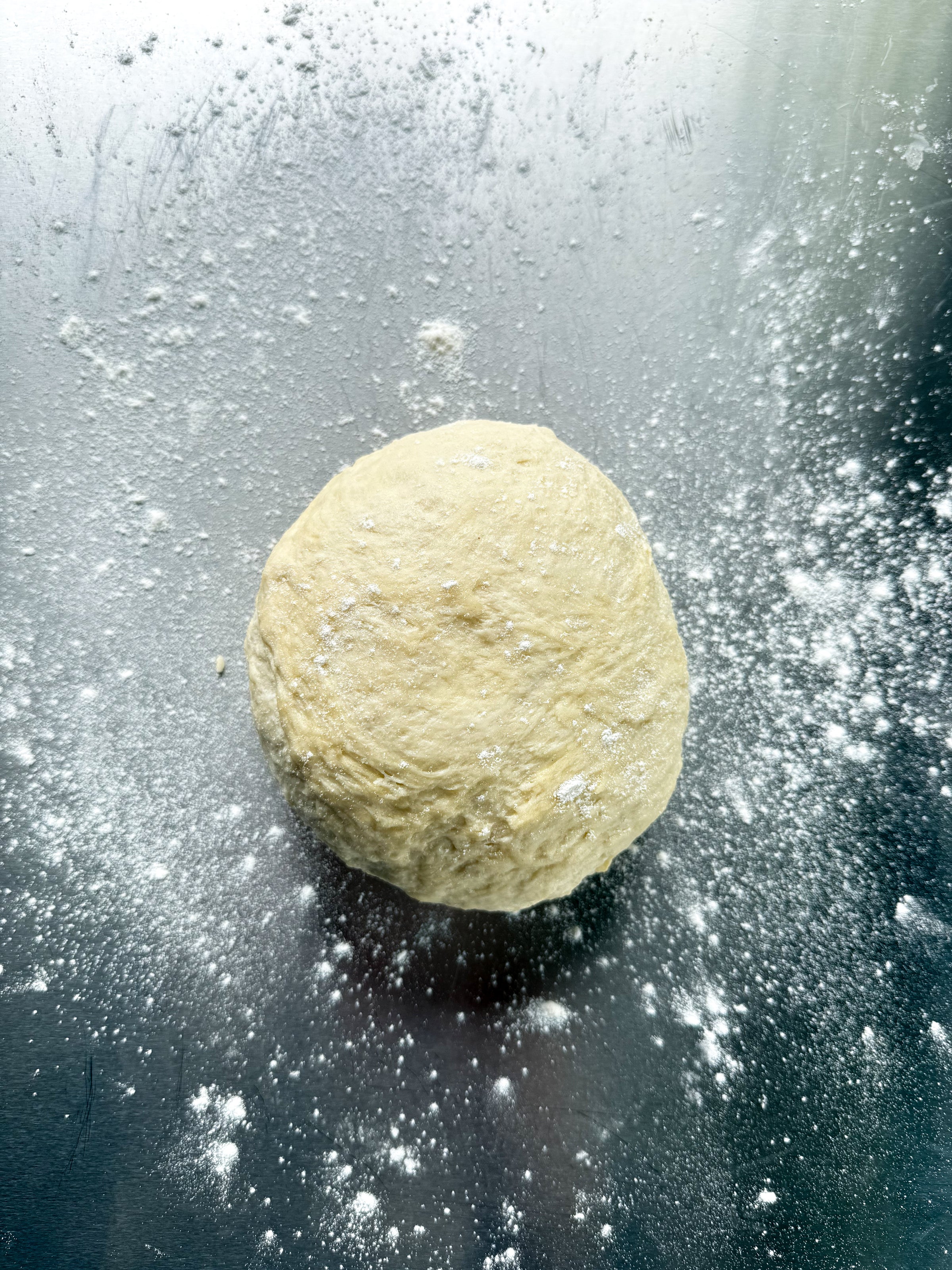 A ball of dough sits on a floured metallic surface, with flour sprinkled around it.