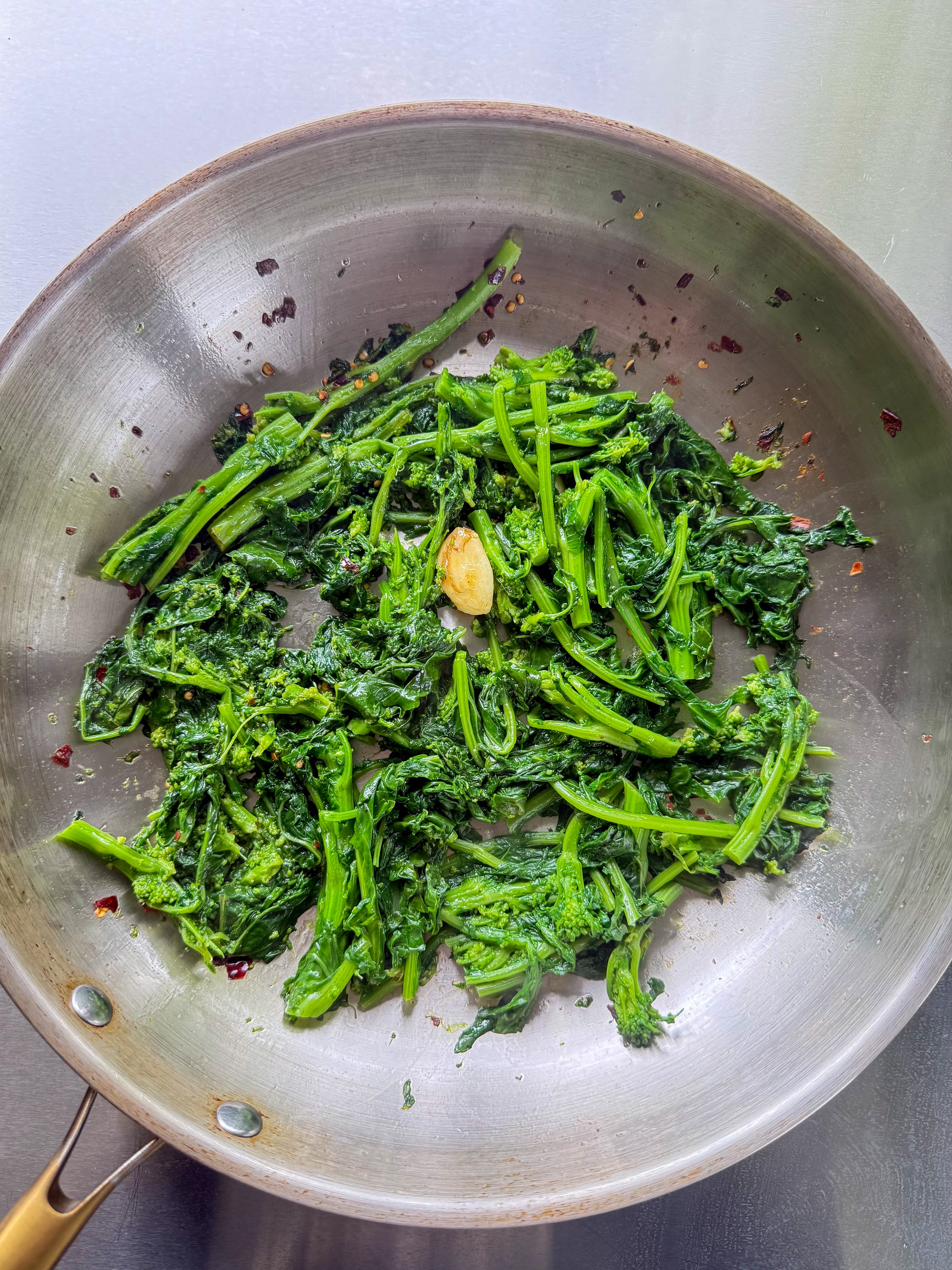 Cooked broccoli rabe with garlic and red pepper flakes in a stainless steel pan on a gray surface. The greens appear sautéed and lightly wilted.