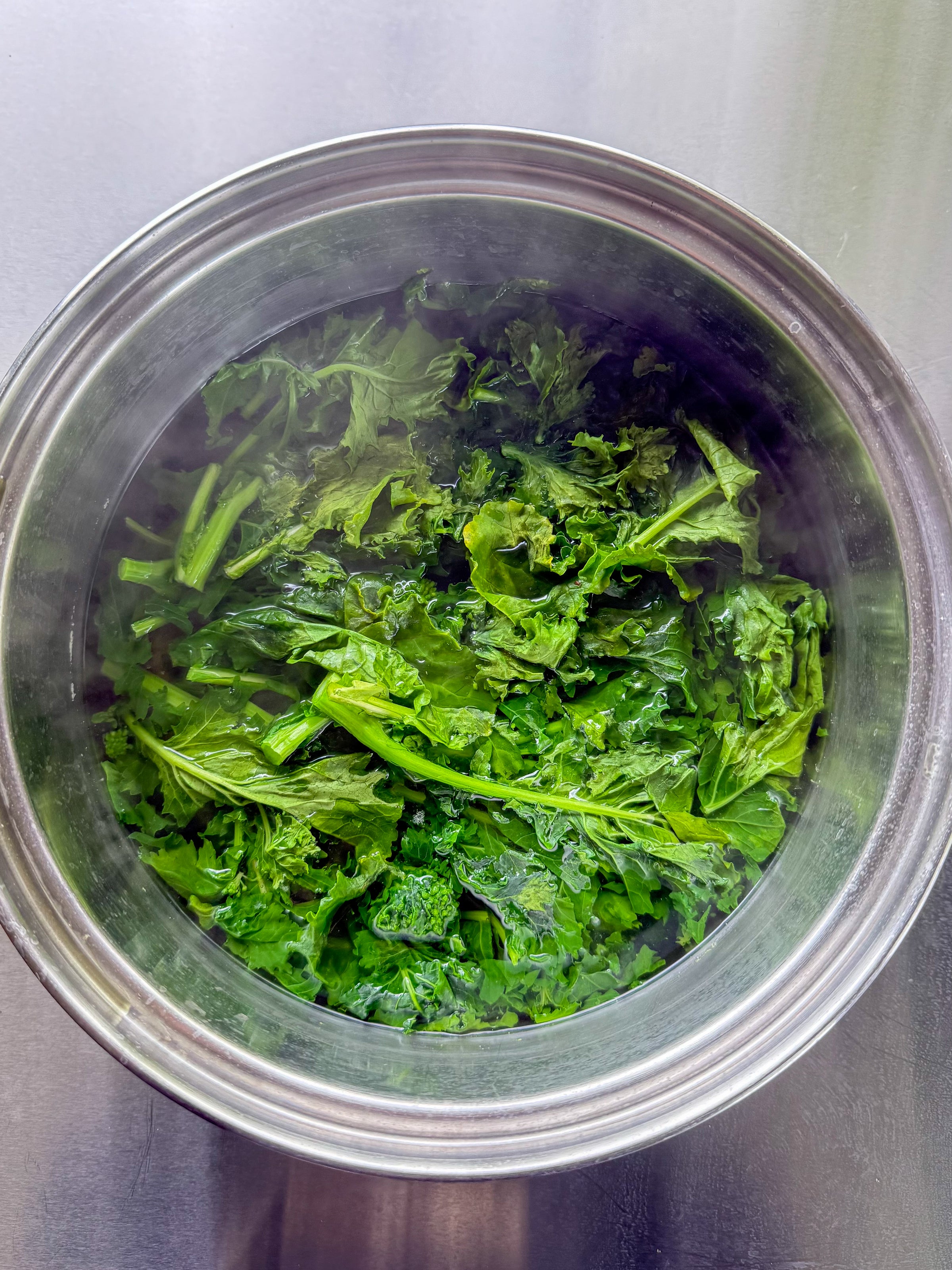 A pot filled with freshly steamed kale sits on a metal surface. Steam is visible rising from the vibrant green leafy vegetables.