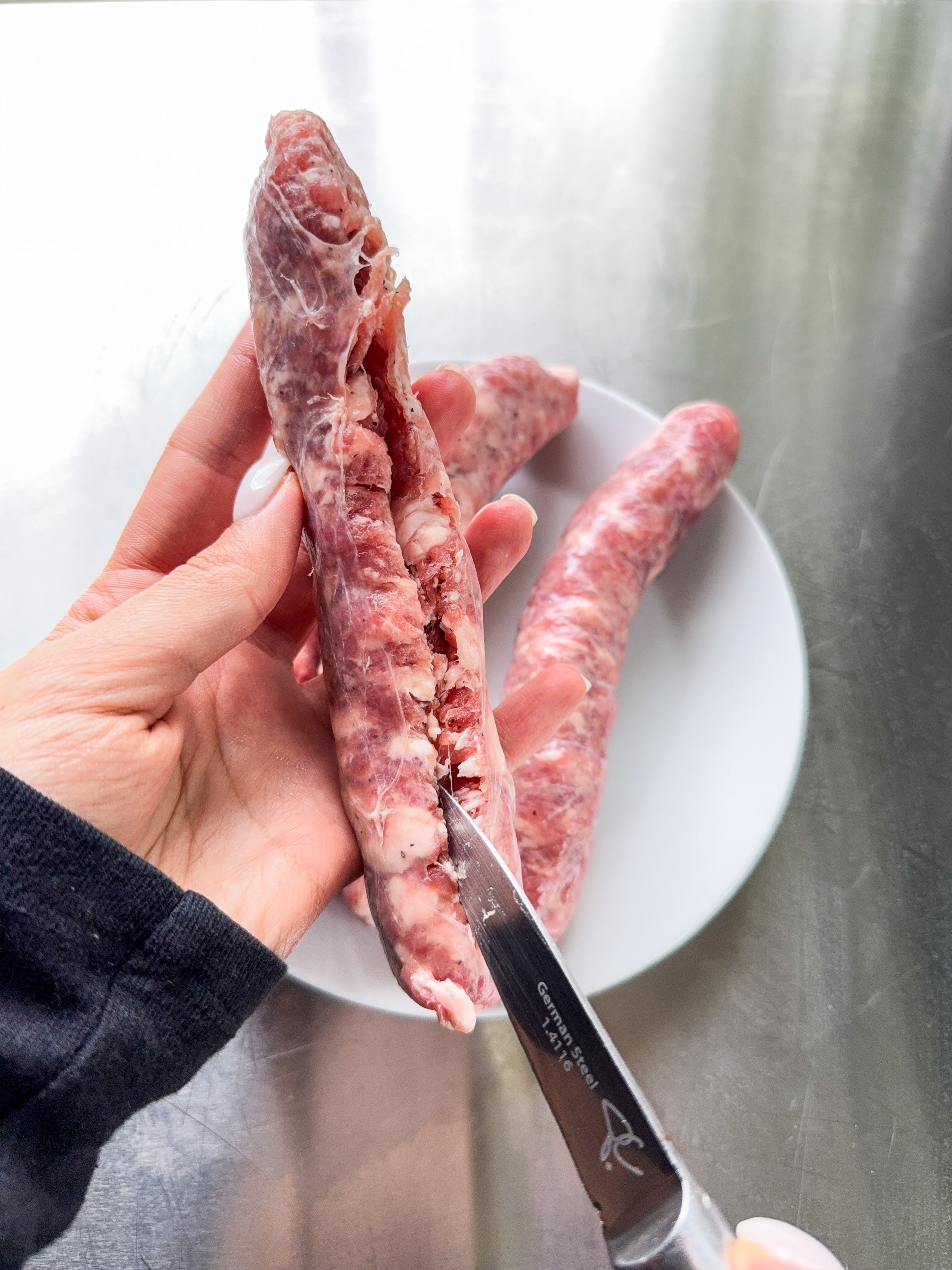 A hand holds a raw turkey neck while using a knife to cut it open; another turkey neck rests on a white plate in the background on a metallic surface.
