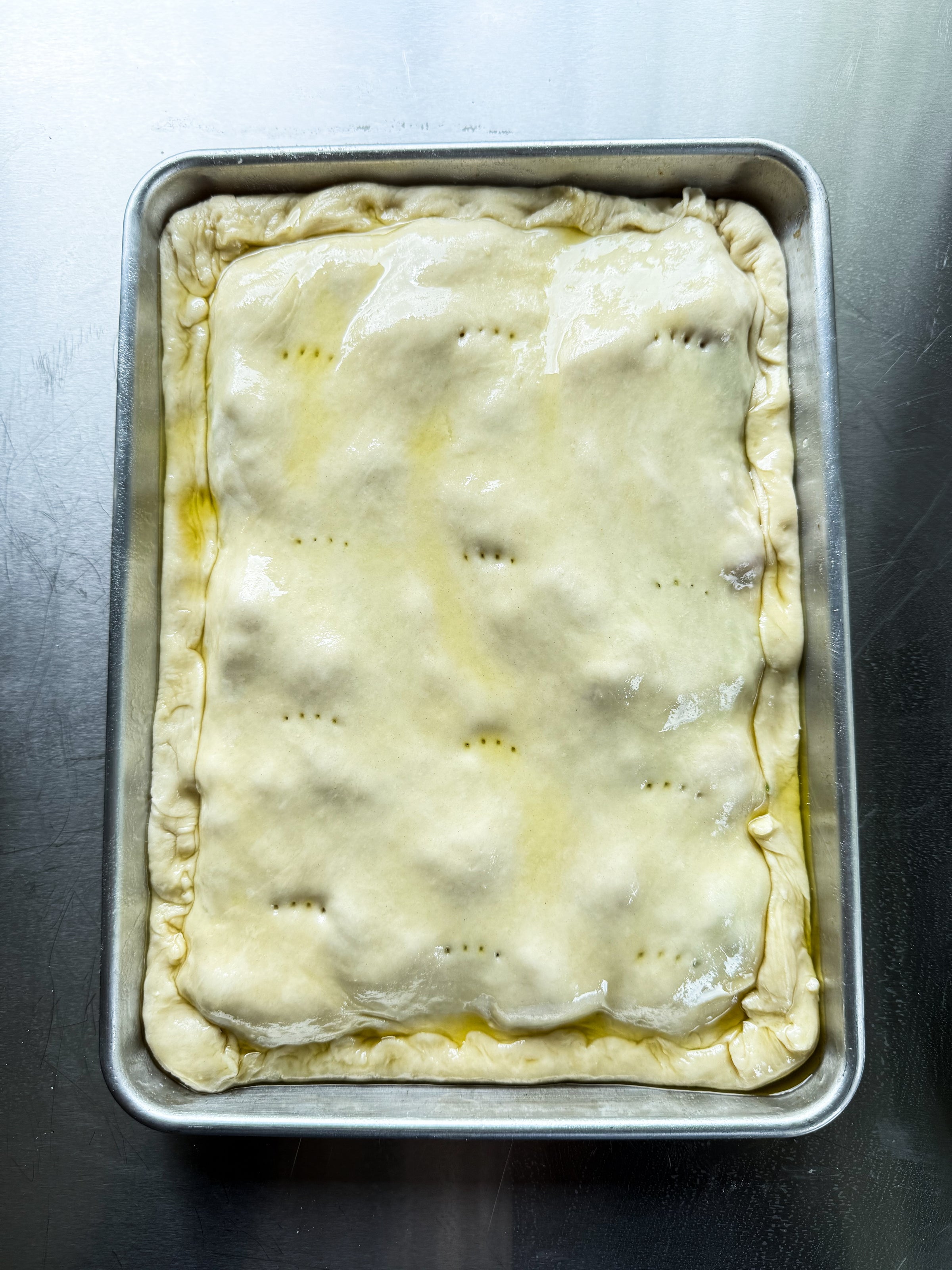 A rectangular unbaked pie in a metal baking pan, with a crimped edge and a shiny, egg-washed top crust that has several small fork holes. The pan rests on a reflective metal surface.