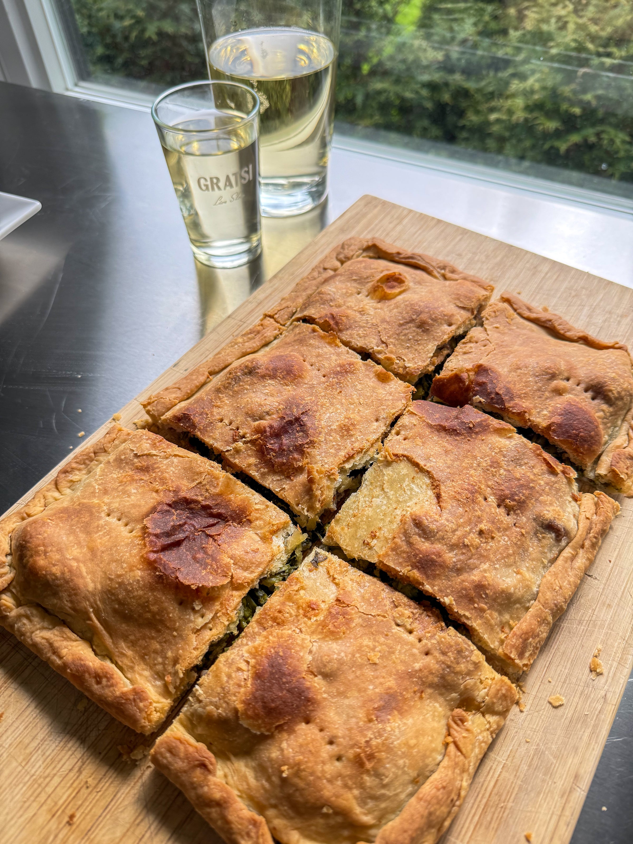 A golden-brown savory pie, cut into six squares, sits on a wooden board next to two glasses of white wine by a window with a green garden view.