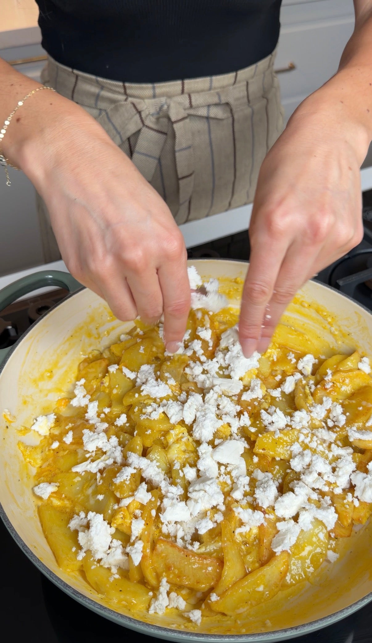 A person crumbles white cheese over a yellow, creamy dish in a pan on the stove. The person is wearing a striped apron and a delicate bracelet.