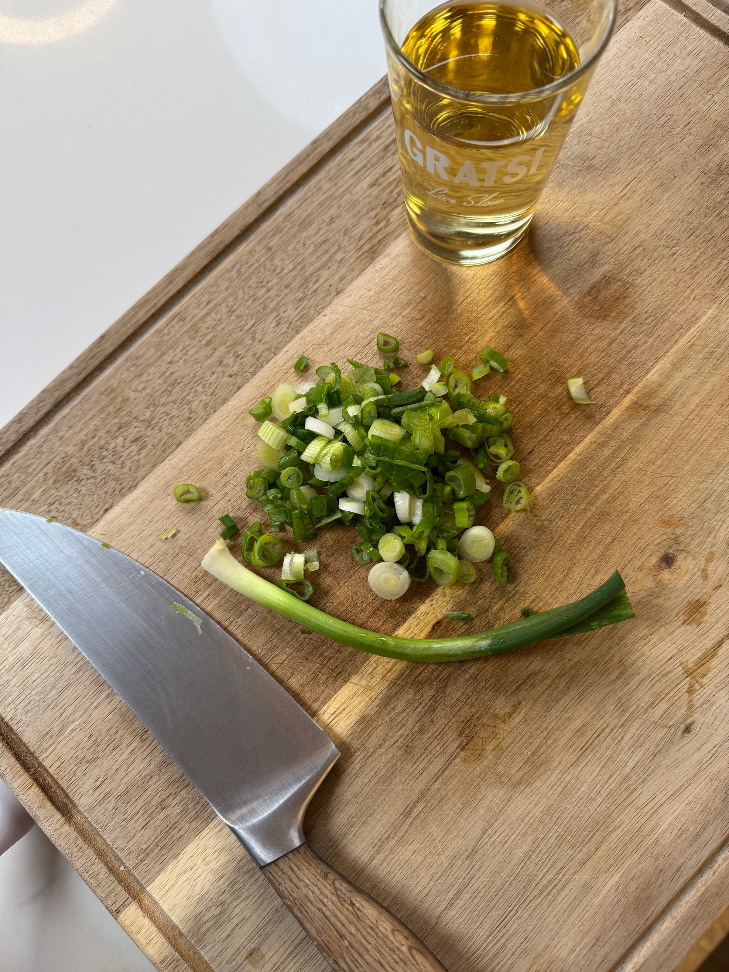 A wooden cutting board with a kitchen knife, chopped green onions, a whole green onion, and a glass of yellow liquid.