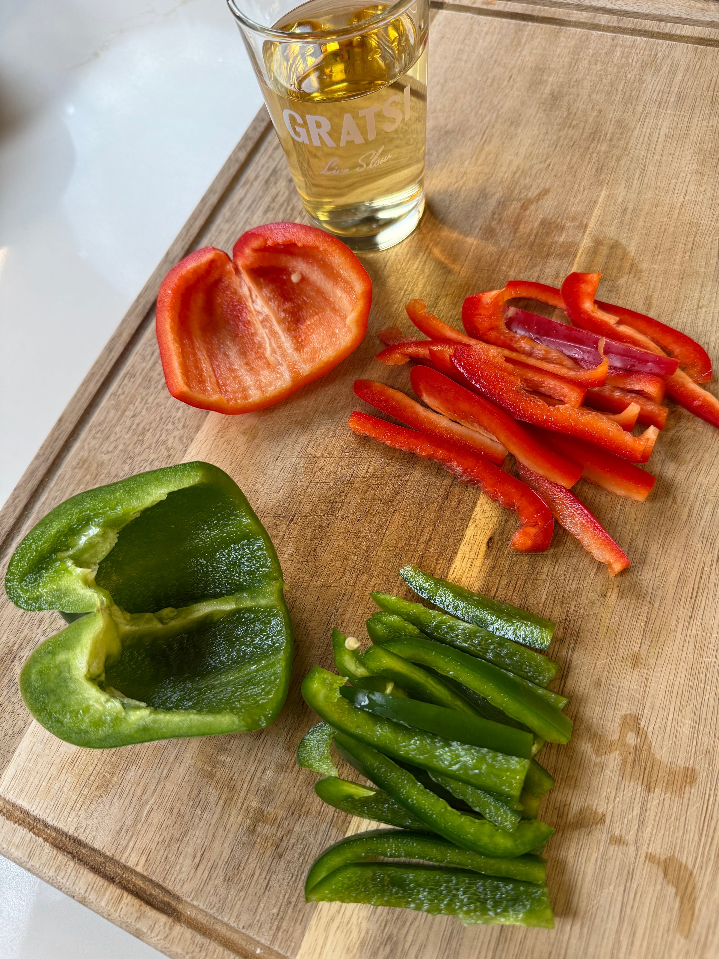 A wooden cutting board with sliced and halved red and green bell peppers, next to a glass of light-colored liquid labeled “GRATSI.” The vegetables are arranged in groups on the board.