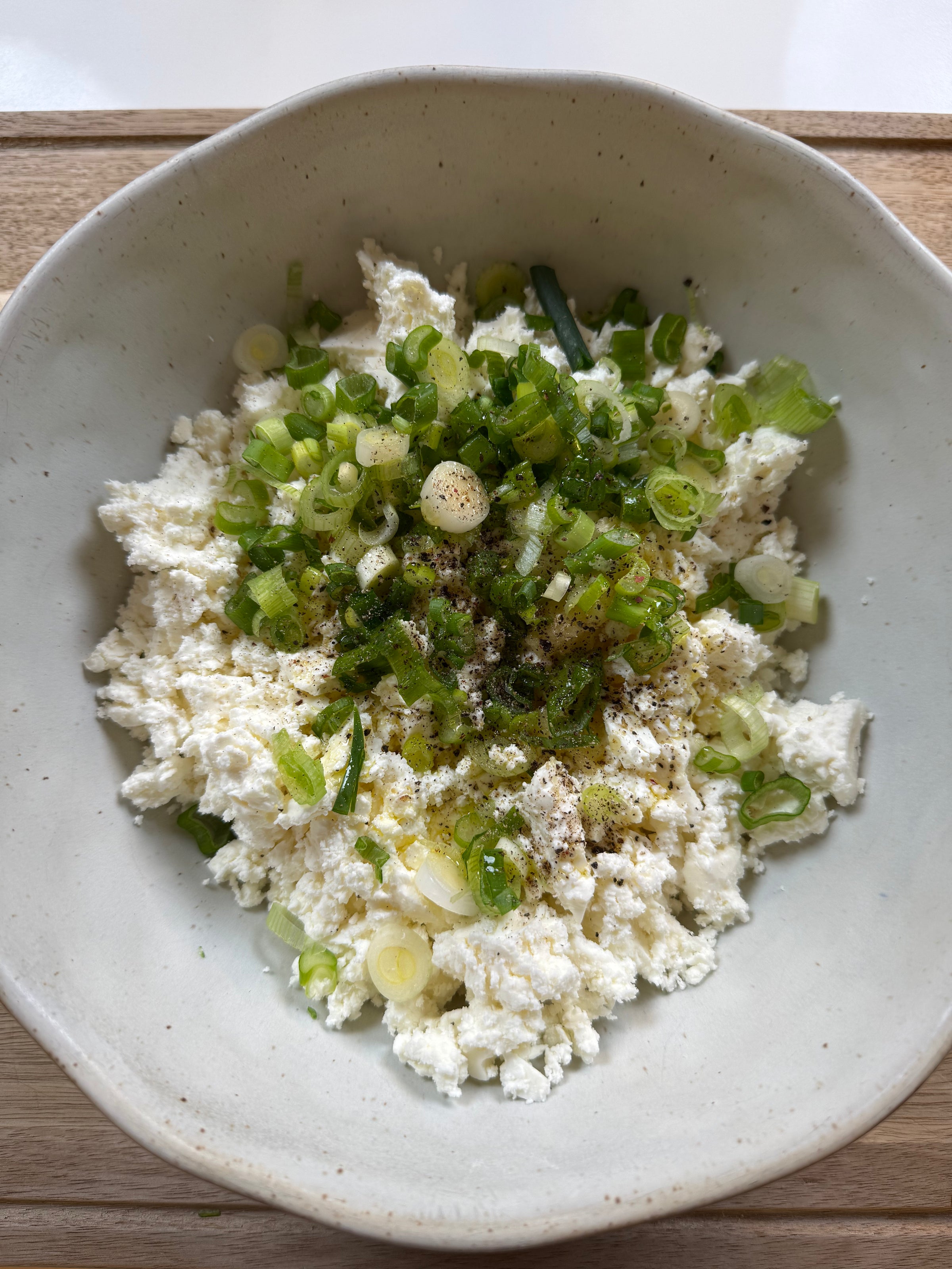 A bowl of crumbled white cheese topped with chopped green onions, cracked black pepper, and seasoning, served in a light-colored ceramic bowl on a wooden surface.