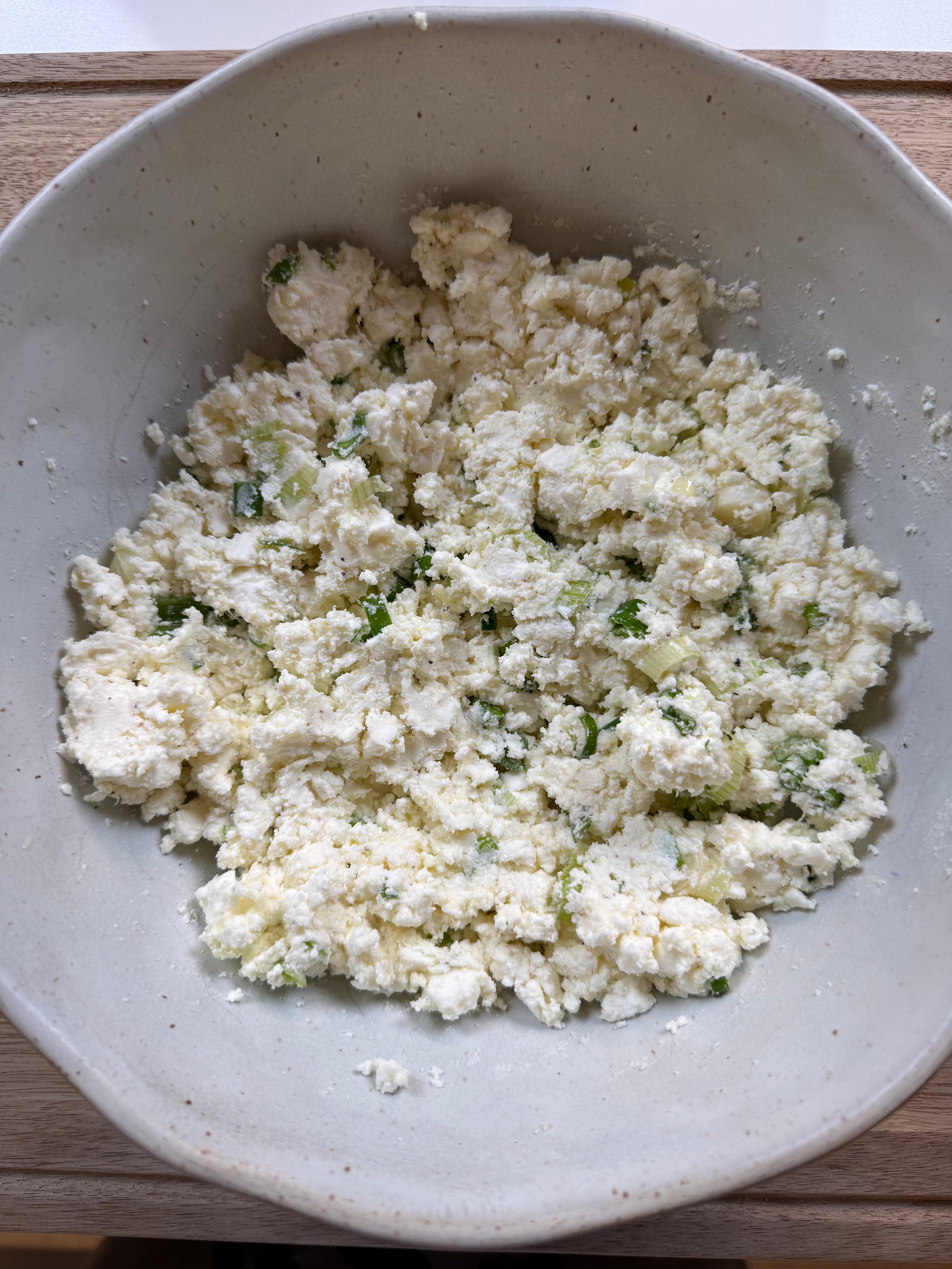 A white ceramic bowl filled with crumbly homemade ricotta cheese mixed with chopped herbs, resting on a wooden surface.