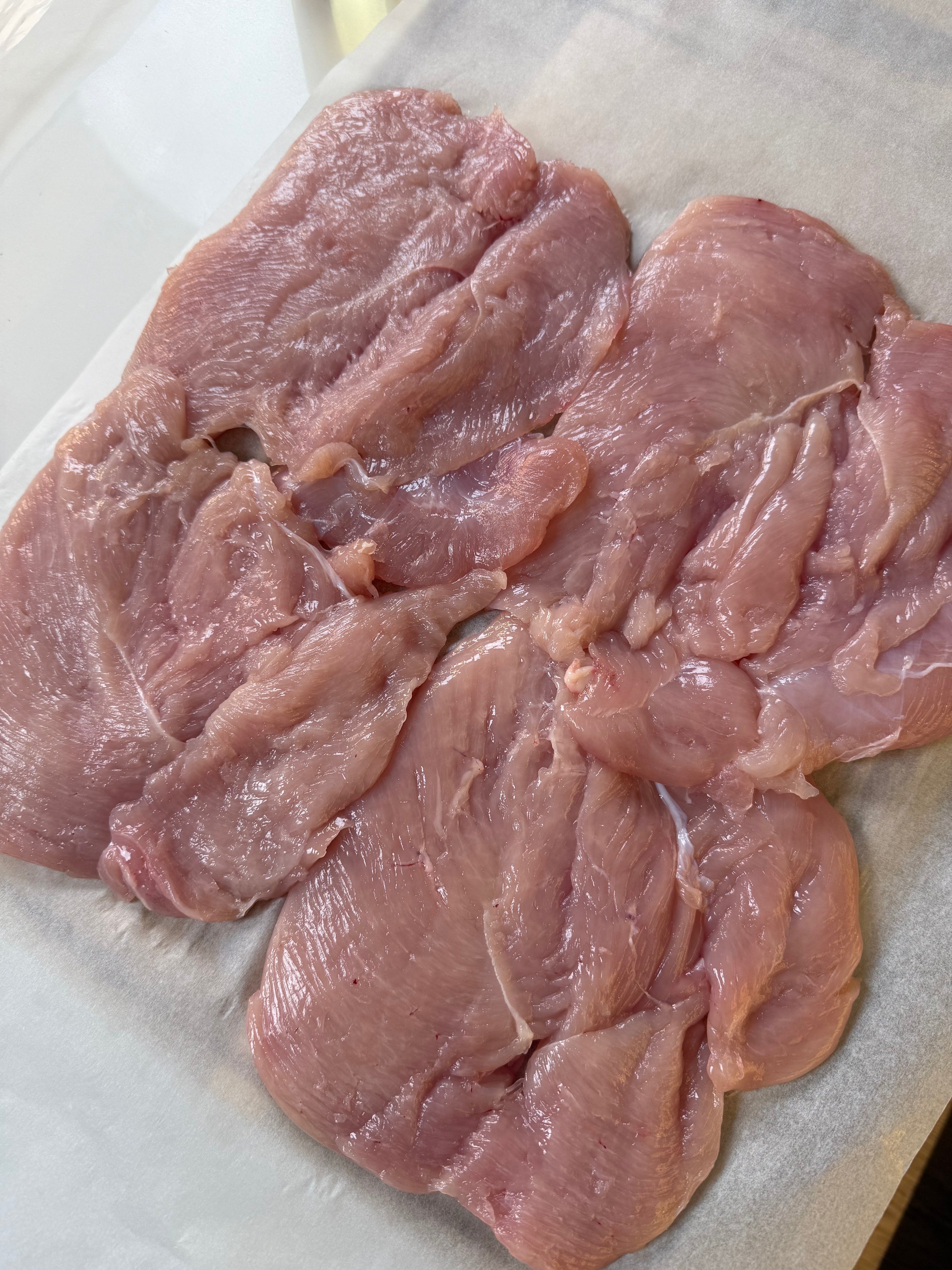 Raw, uncooked chicken breasts are laid flat on a sheet of parchment paper, ready for preparation. The meat appears fresh and pink, with visible muscle texture and slight marbling.