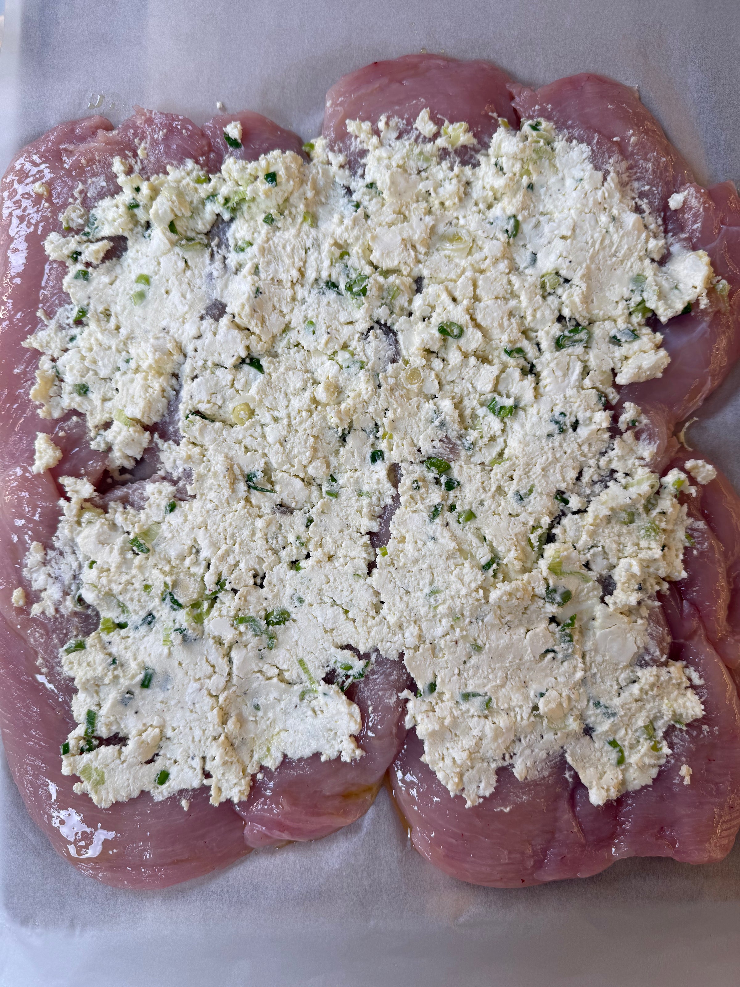 Raw, butterflied chicken breast laid flat on parchment paper, topped with a spread of herbed ricotta cheese mixture containing green herbs or chives.