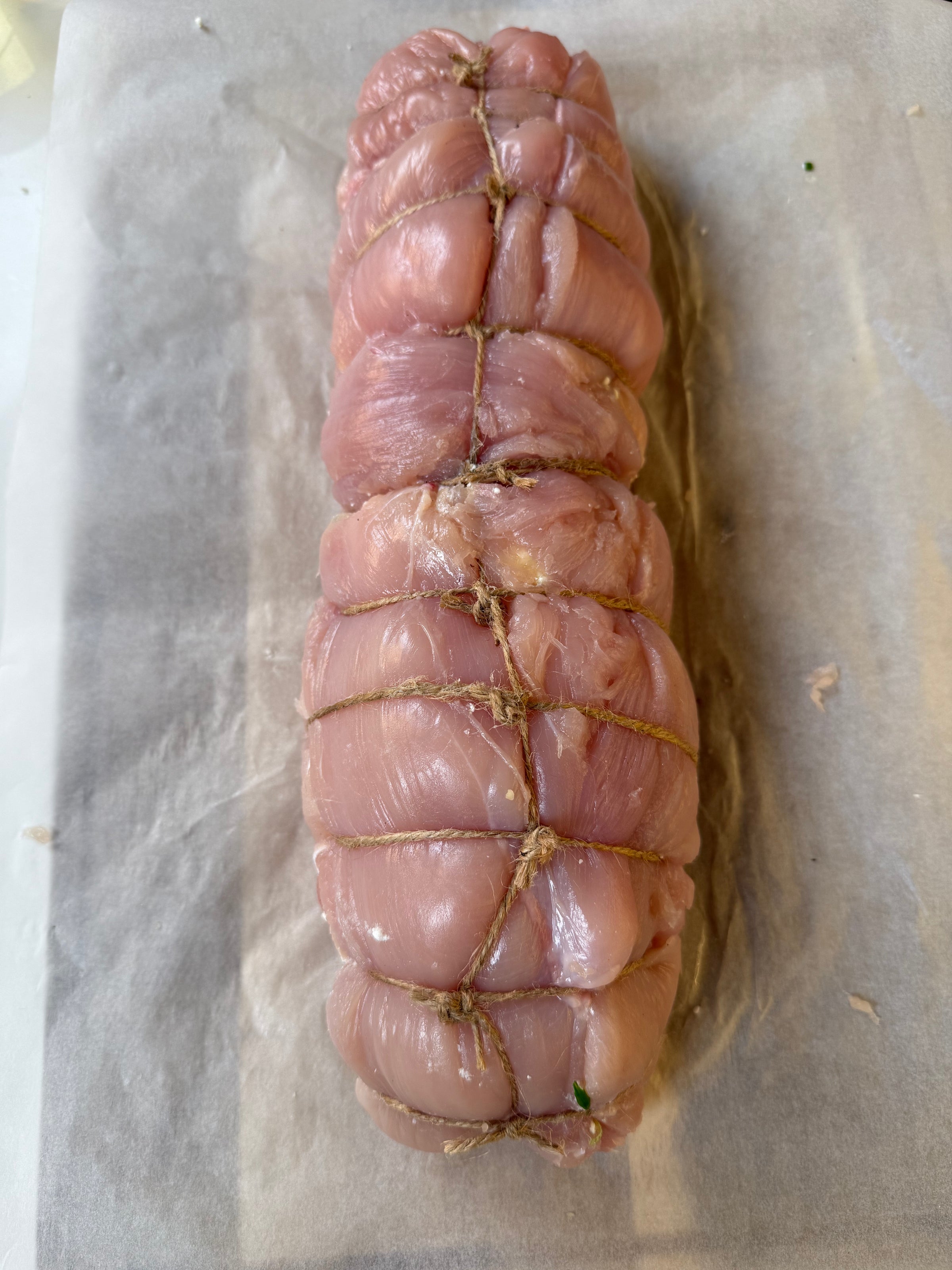 A raw, boneless piece of meat is tightly tied with kitchen twine in a roast shape, resting on a sheet of parchment paper.