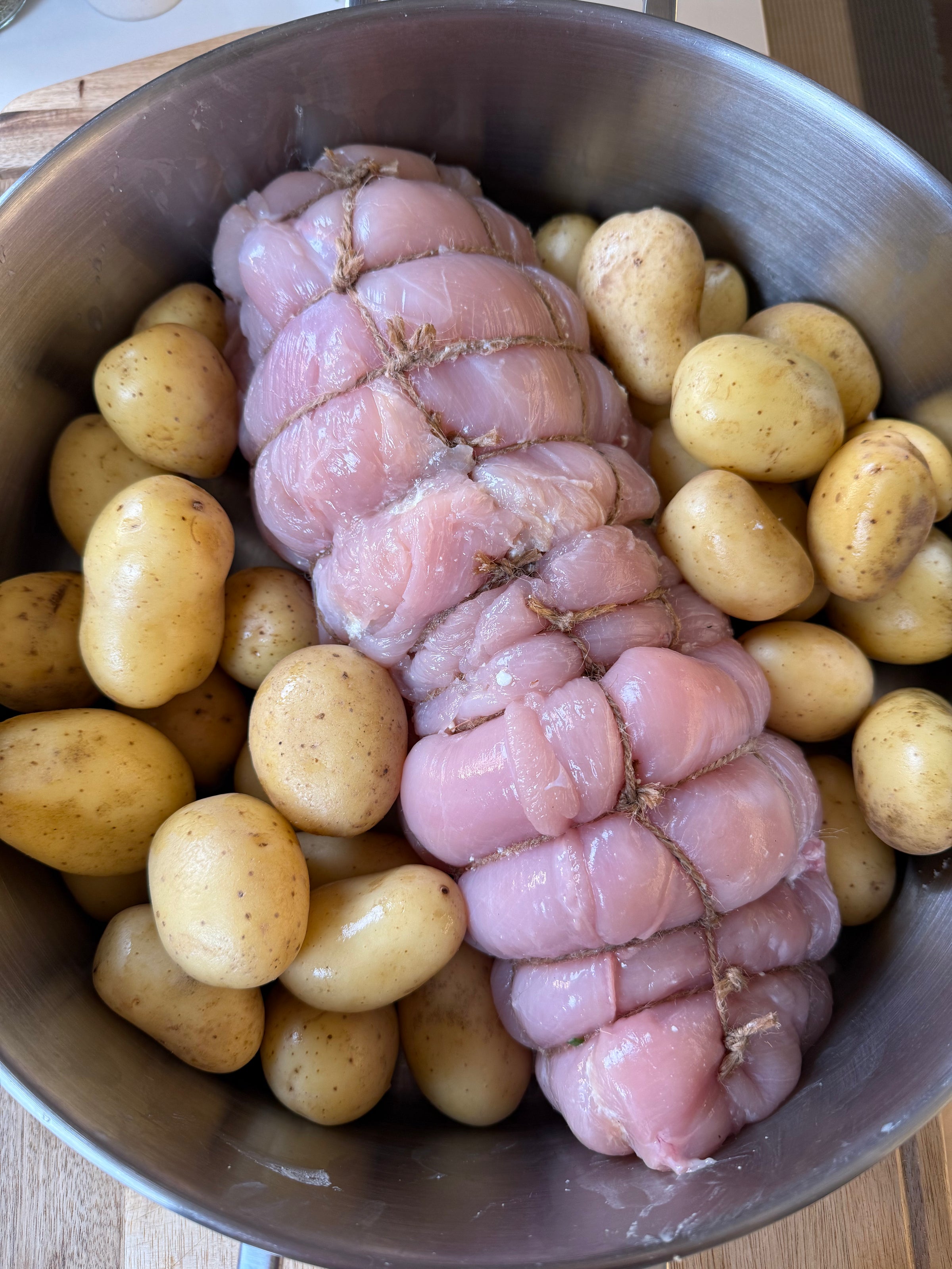 A large, tied and trussed raw meat roast surrounded by small, unpeeled potatoes in a metal bowl, ready for cooking.