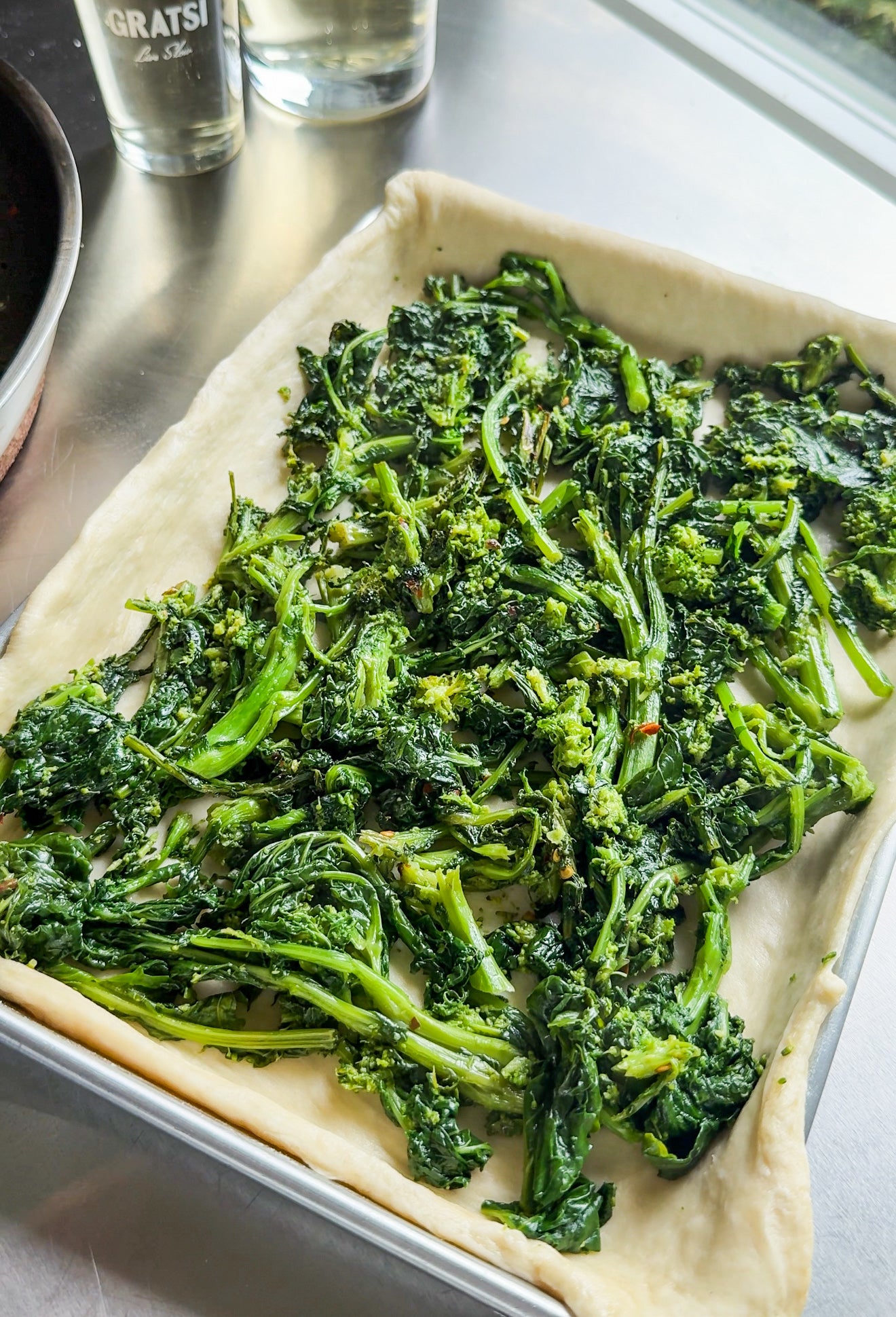 A baking sheet with raw pizza dough topped with sautéed broccoli rabe sits on a metal countertop near a window, with two glass bottles in the background.
