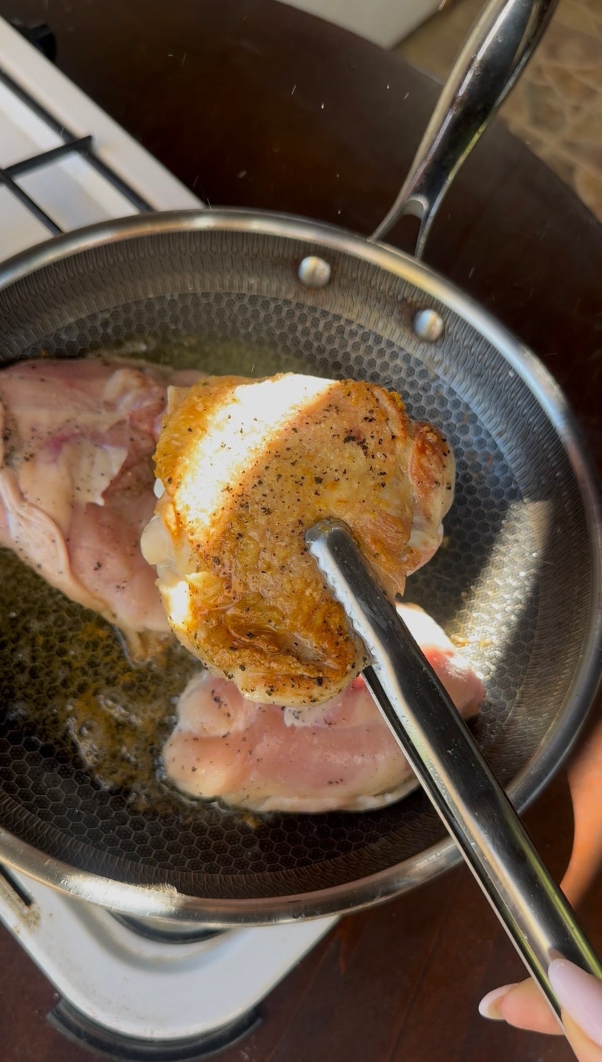 A person uses metal tongs to sear chicken thighs in a frying pan; one piece shows crispy golden-brown skin while others are still raw.
