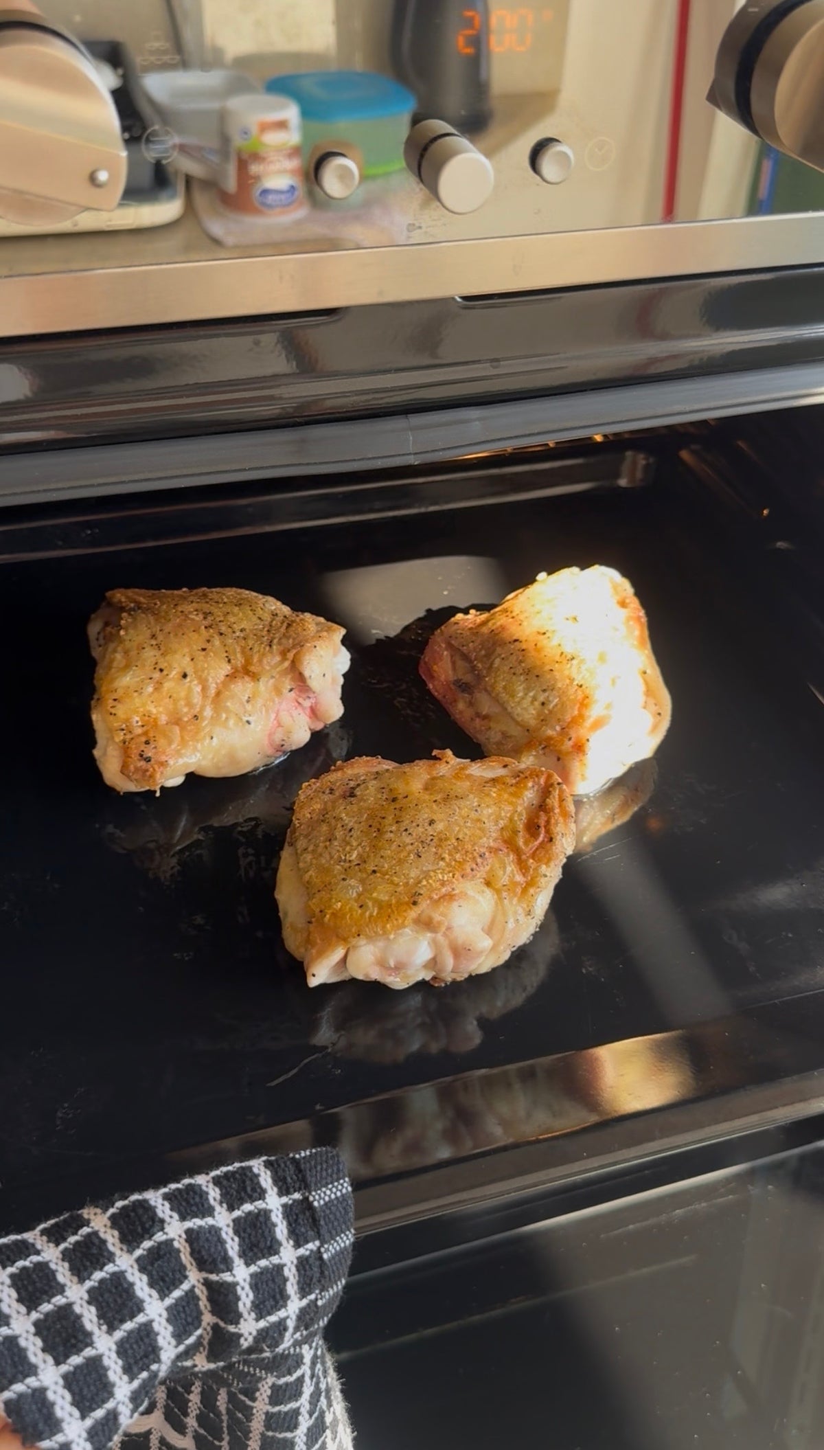 Three seasoned chicken thighs cooking on a black oven tray, with sunlight shining on them. A checkered kitchen towel is visible in the lower left corner.