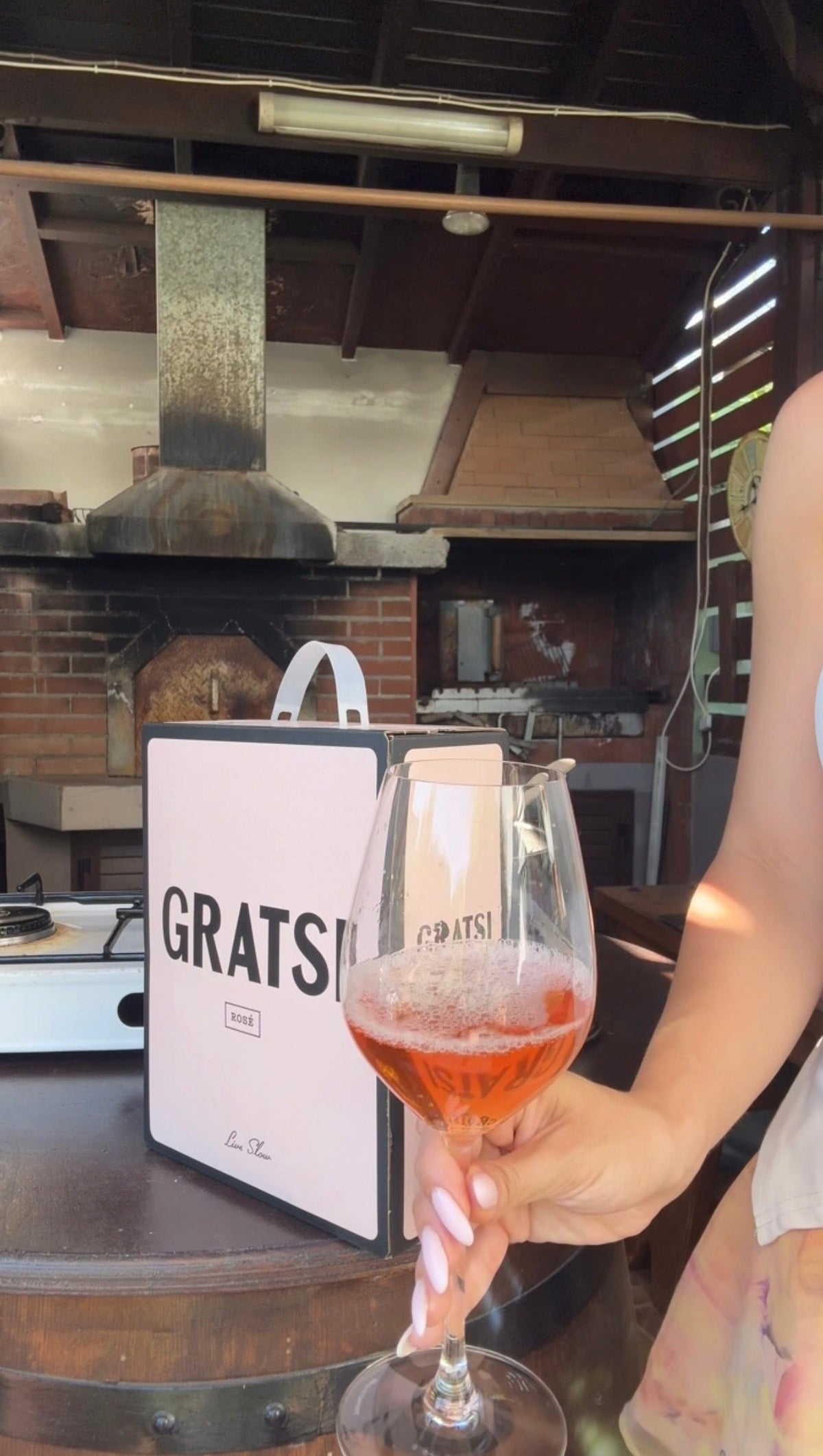 A person holds a glass of rosé wine in front of a GRATS! wine box, with a brick pizza oven and outdoor kitchen setting in the background.