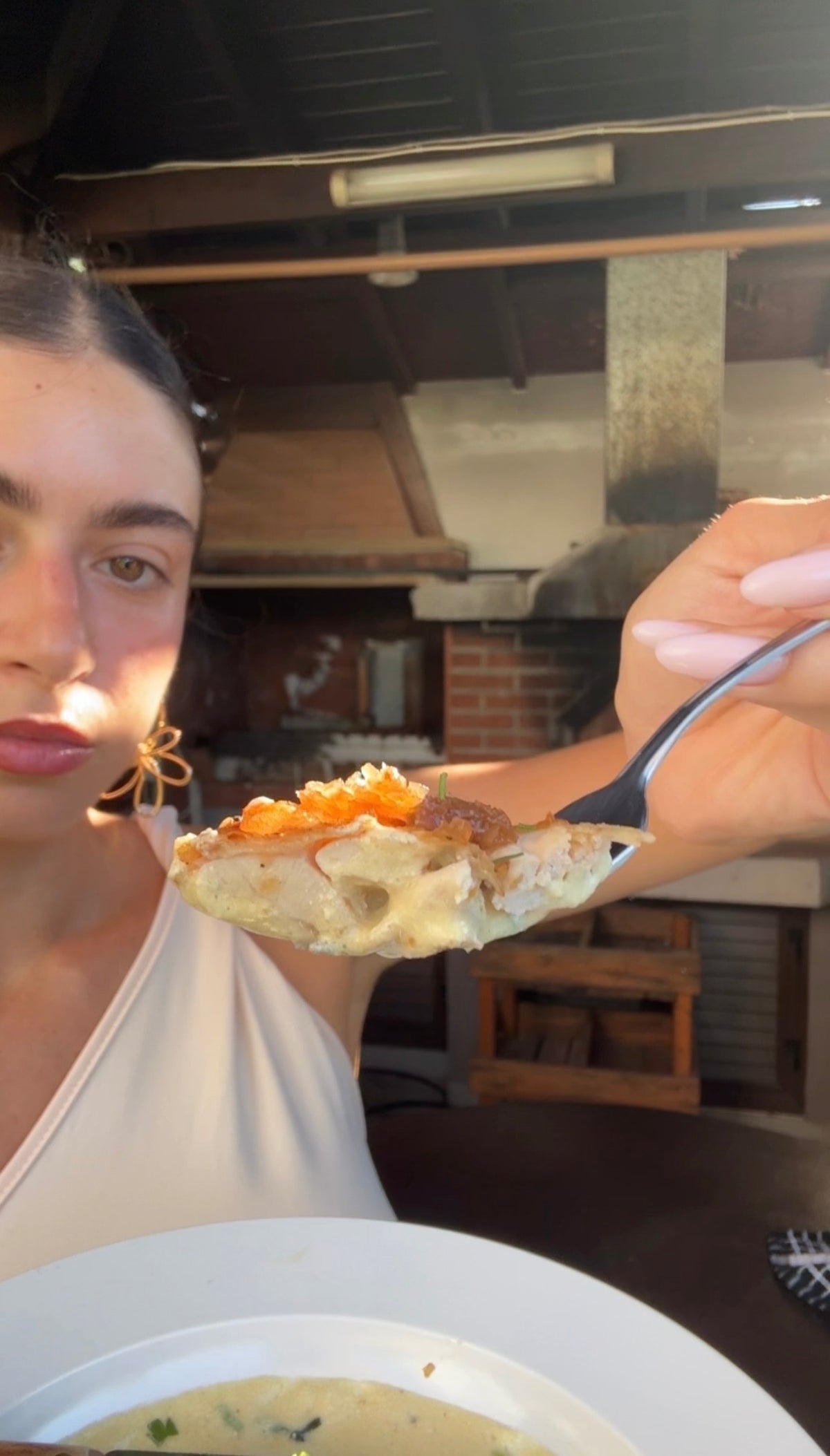 A woman holds a spoon close to the camera with a bite of food topped with orange garnish, while looking at it. A brick oven and wooden furniture are in the background.