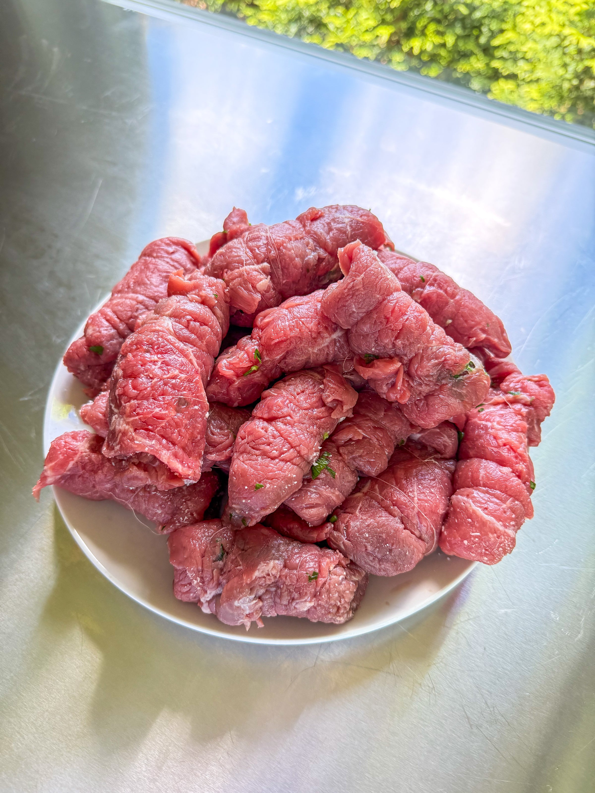 A plate piled with raw beef rolls stuffed with herbs, placed on a shiny metal surface near a window with greenery visible outside.