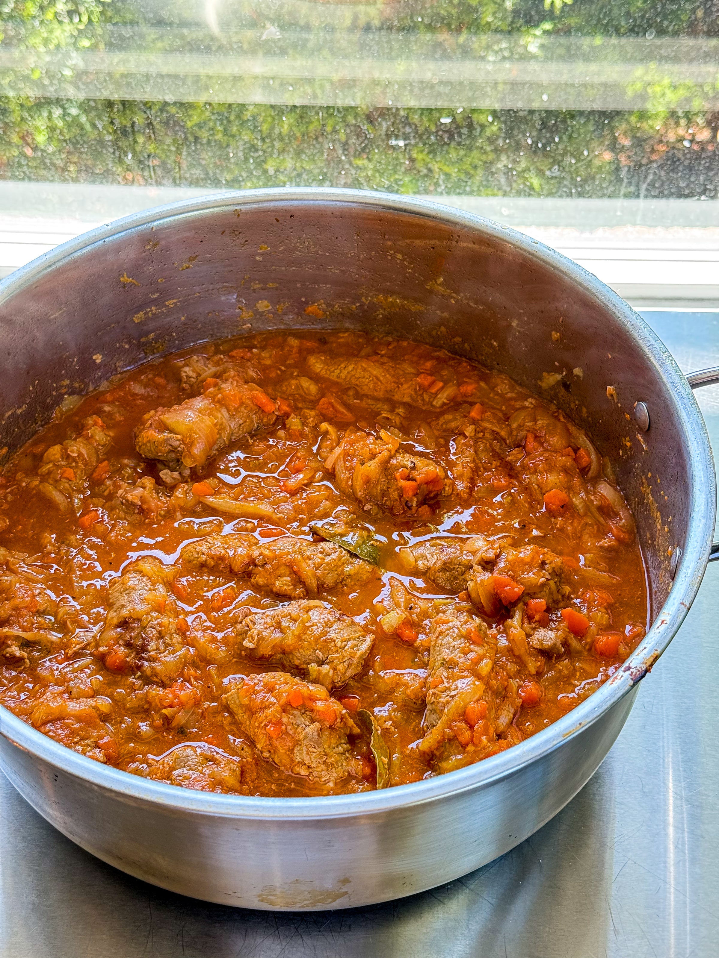 A large metal pot filled with a hearty stew featuring pieces of meat in a thick, chunky tomato-based sauce, sitting on a countertop by a sunlit window.
