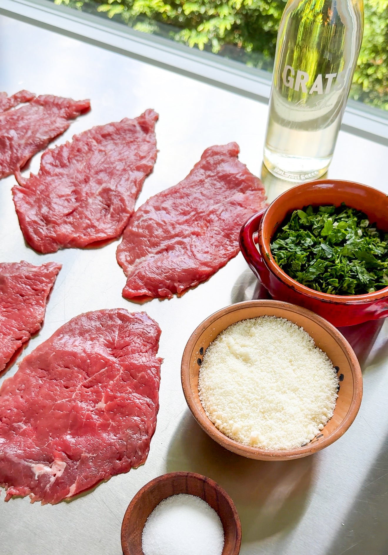 Thin slices of raw beef, chopped parsley in a red bowl, grated cheese in a brown bowl, a small bowl of salt, and a bottle of white wine are arranged on a metal surface near a window.