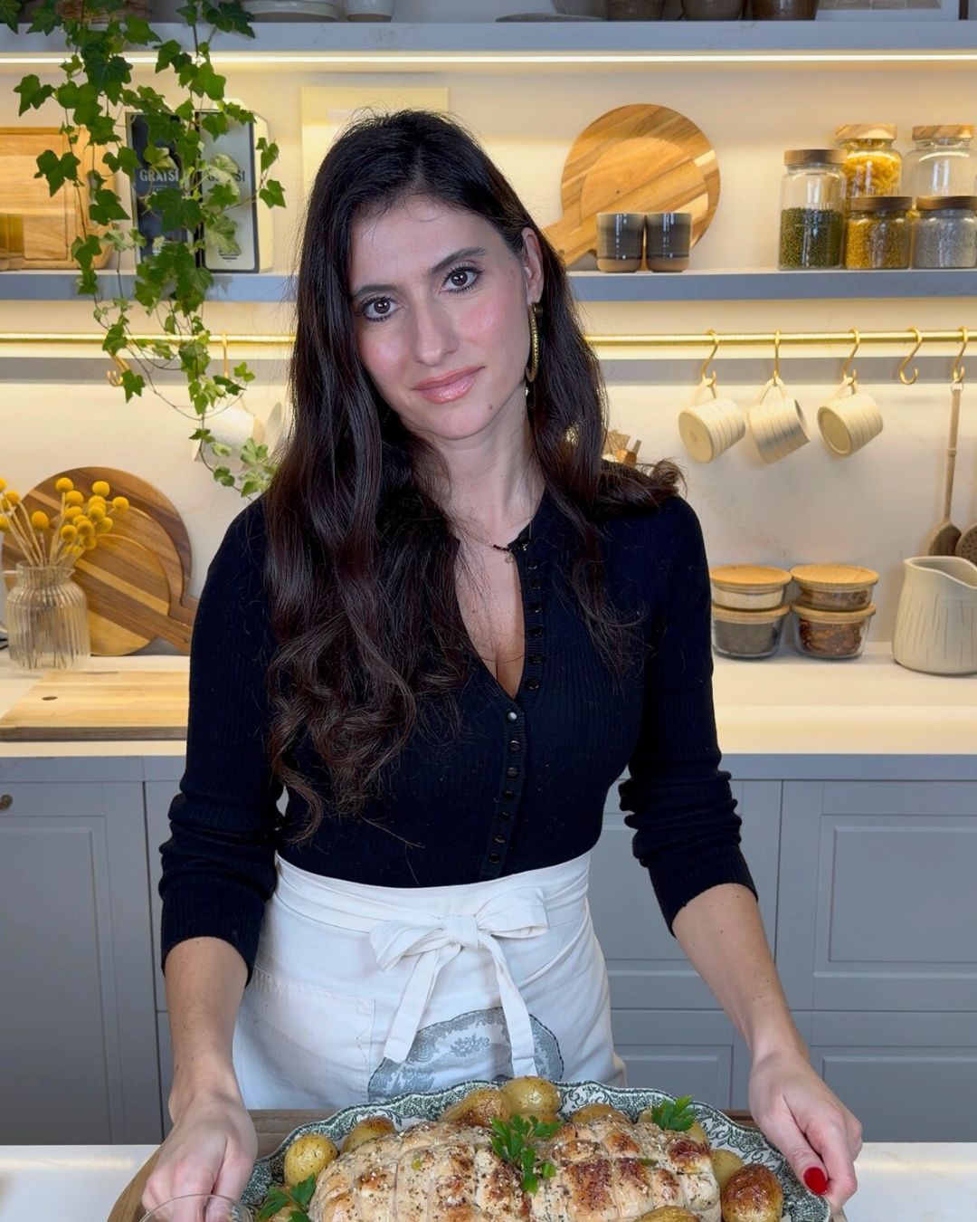 A woman with long dark hair, wearing a black top and a cream apron, stands in a modern kitchen holding a platter of rolled stuffed chicken and roasted potatoes, with shelves and hanging cups in the background.
