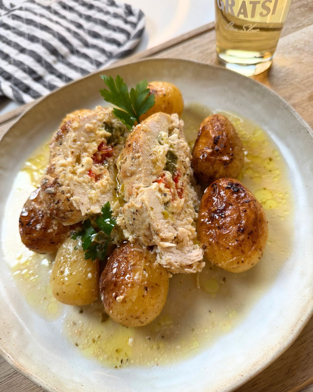 A plate with stuffed chicken breast, garnished with parsley, and served with roasted baby potatoes in a light herb sauce. A glass of drink and a striped napkin are in the background.