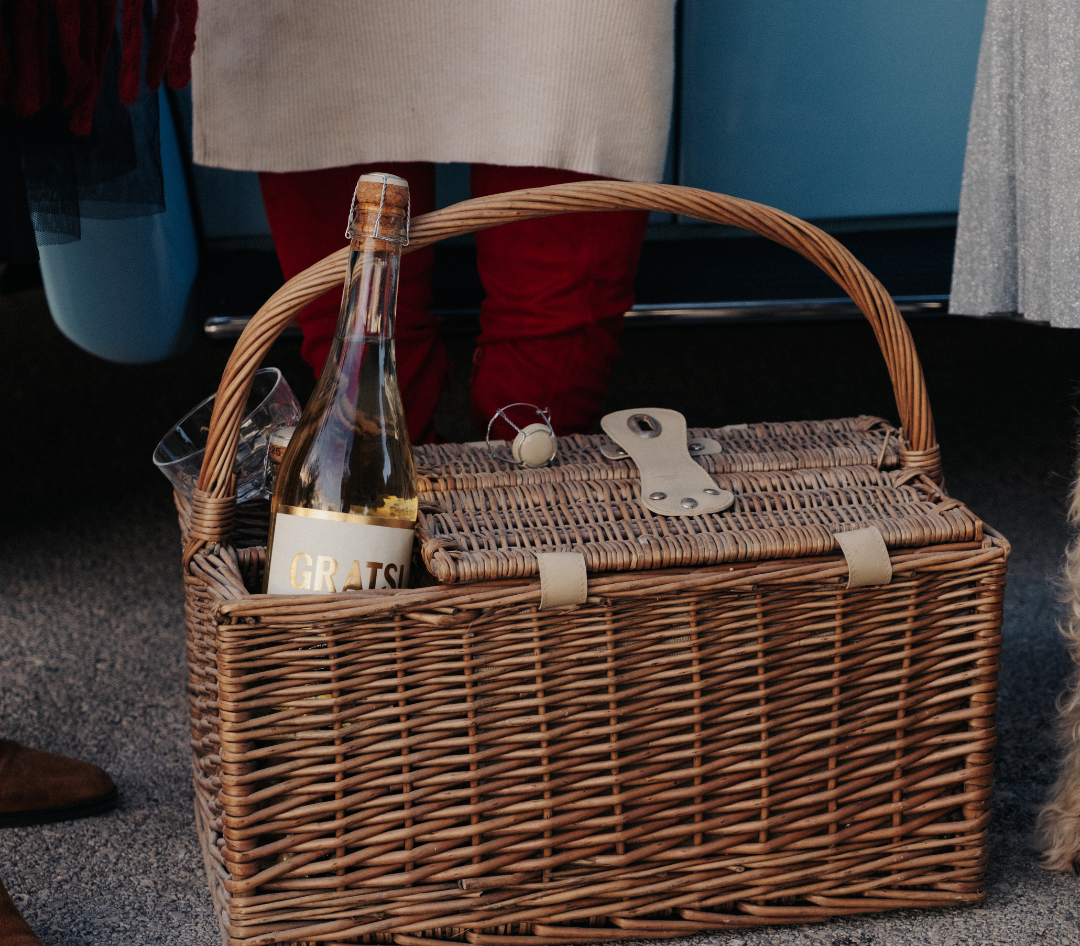 A wicker picnic basket with a bottle of white wine, a corkscrew, and a glass sits on the ground next to people wearing red and beige clothing.