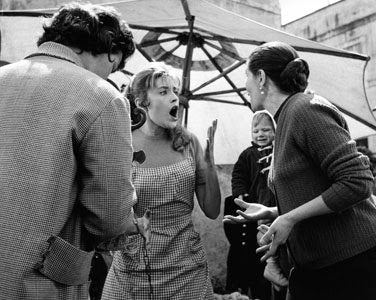 A woman in a checkered dress stands outdoors, appearing shocked with her mouth open and hands raised. She faces another woman gesturing animatedly; a person with headphones and a microphone is nearby. An umbrella and child are in the background.