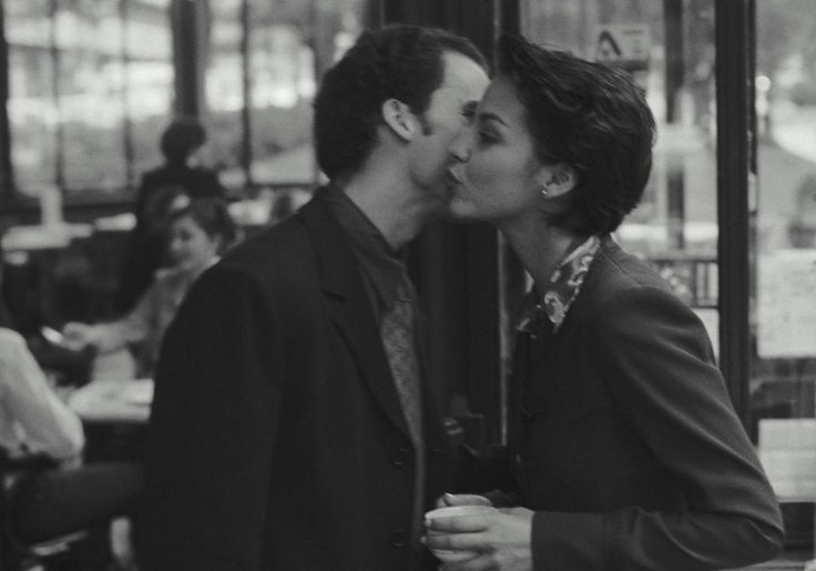 A man and a woman greet each other with a cheek kiss in a busy café. The image is black and white, and other people can be seen sitting and talking in the blurred background.