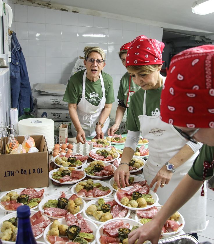 Three women in red bandanas and green shirts prepare plates of assorted food, including cold cuts and vegetables, in a kitchen. One woman arranges food while another looks up and speaks. The kitchen has a white tiled wall.