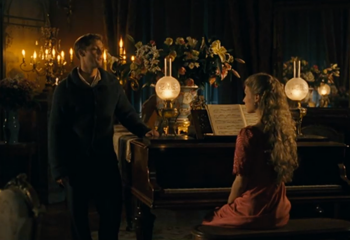 A man stands beside a grand piano where a woman in a pink dress sits, facing sheet music. The room is warmly lit by candles and lamps, and is decorated with flowers and dark drapes.