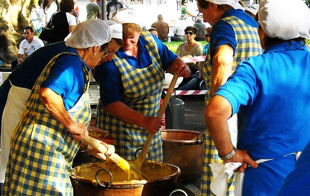 Four women wearing blue shirts, yellow-checked aprons, and hairnets stir large pots with wooden paddles outdoors, surrounded by onlookers at what appears to be a public event or festival.