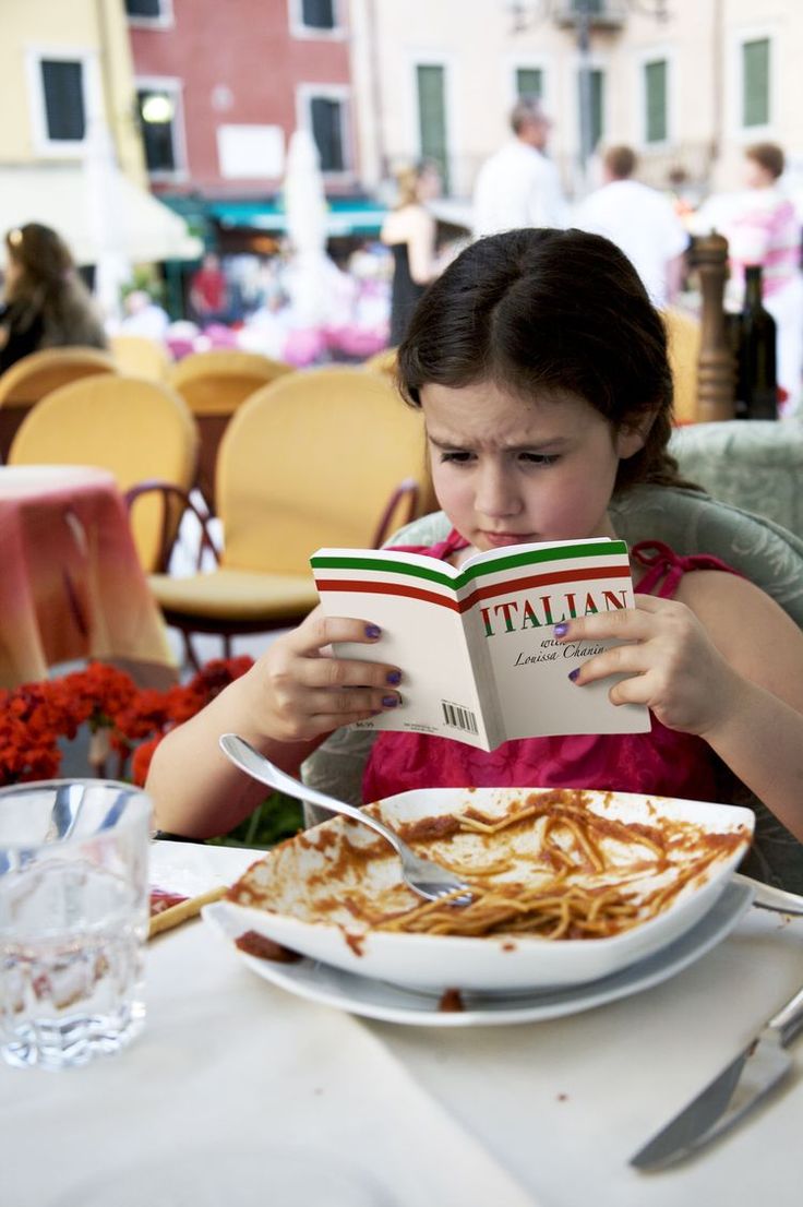 A young girl sits at an outdoor restaurant table with a messy plate of spaghetti, intently reading a small Italian language book. People and colorful chairs fill the background.