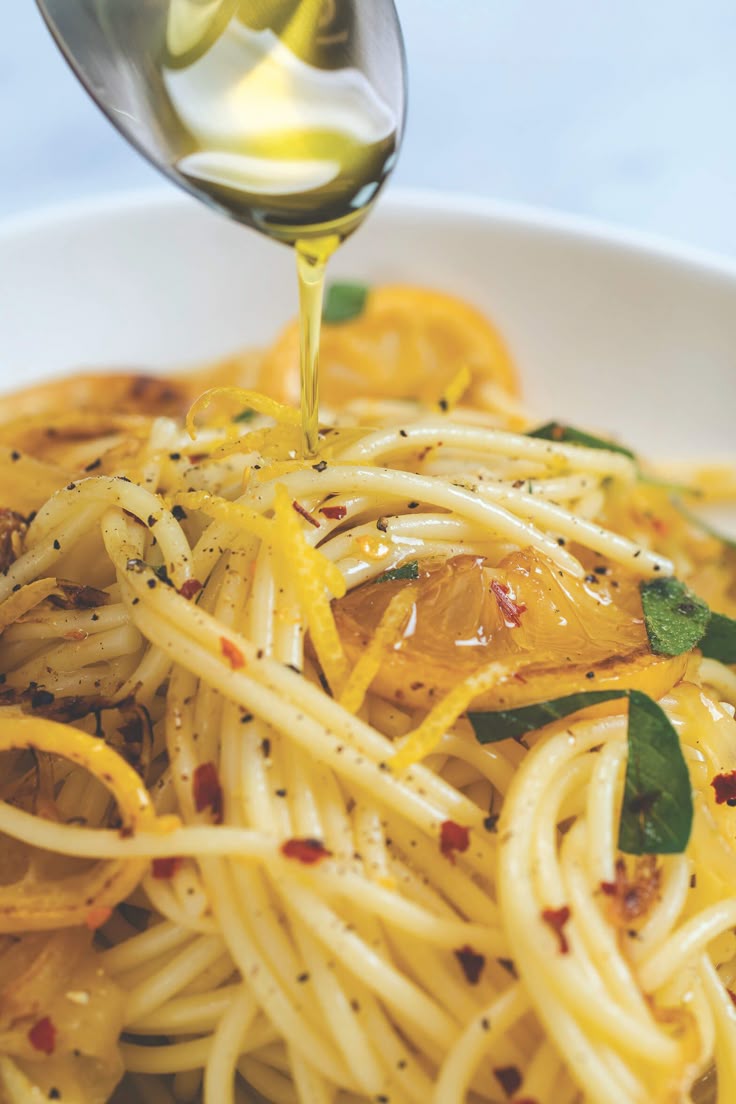 Close-up of cooked spaghetti with lemon slices, fresh herbs, and red pepper flakes, as olive oil is being drizzled from a spoon over the pasta in a white bowl.