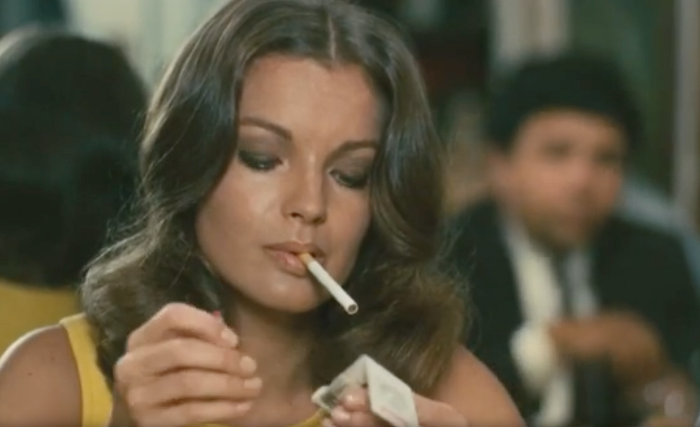 A woman with brown hair sits indoors, holding a cigarette in her mouth while opening a pack. She appears focused on her task. A blurred man sits in the background.
