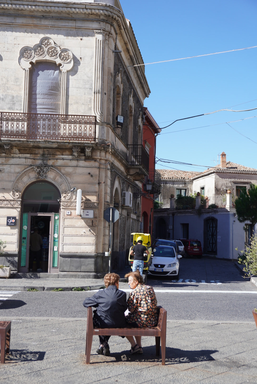 Two elderly women sit on a bench talking in a sunny street in front of an old stone building with ornate balconies. A few cars are parked nearby, and a bright yellow scooter is visible in the background.