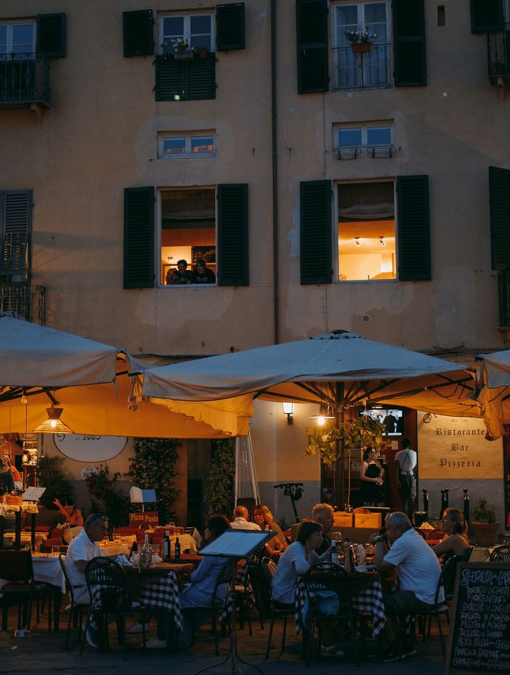 People dine outdoors under large umbrellas at an Italian restaurant in the evening. Warm lights illuminate the terrace and upper floor windows, creating a cozy, lively atmosphere against a historic building facade.
