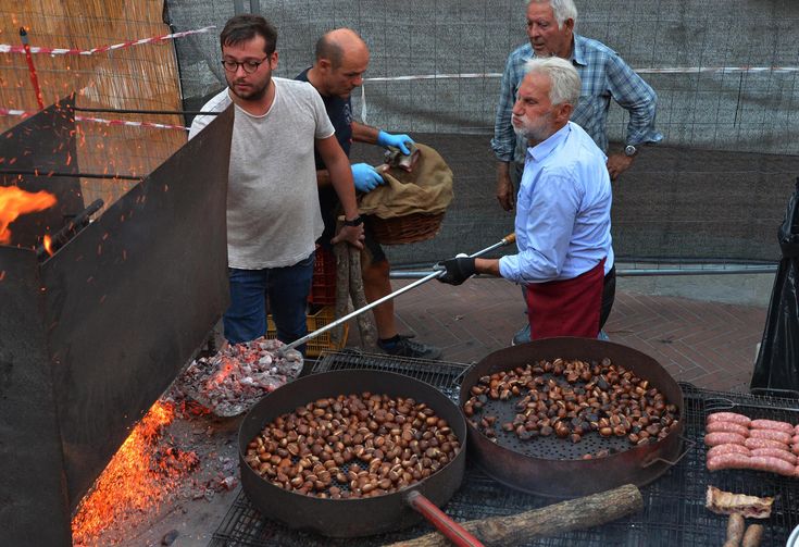 Four men are roasting chestnuts and sausages over open flames at an outdoor event. Two large pans filled with chestnuts are over a grill, and one man tends the fire while others assist. A festive, communal atmosphere is visible.