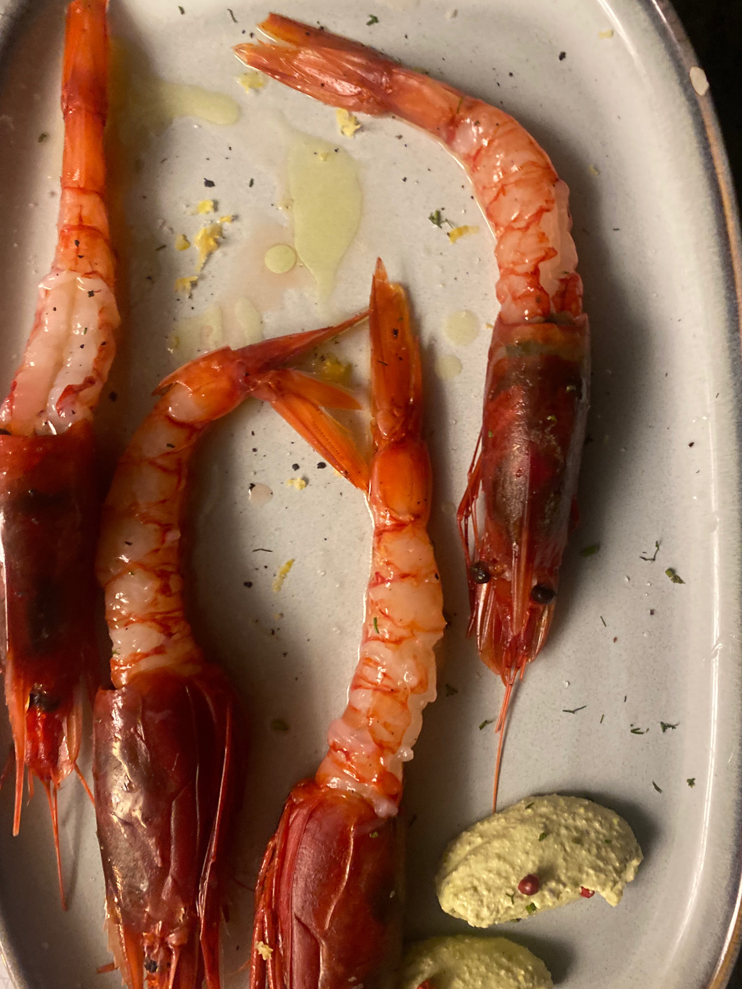 Three raw red prawns arranged on a white plate with a drizzle of oil, herbs, and two dollops of green sauce.