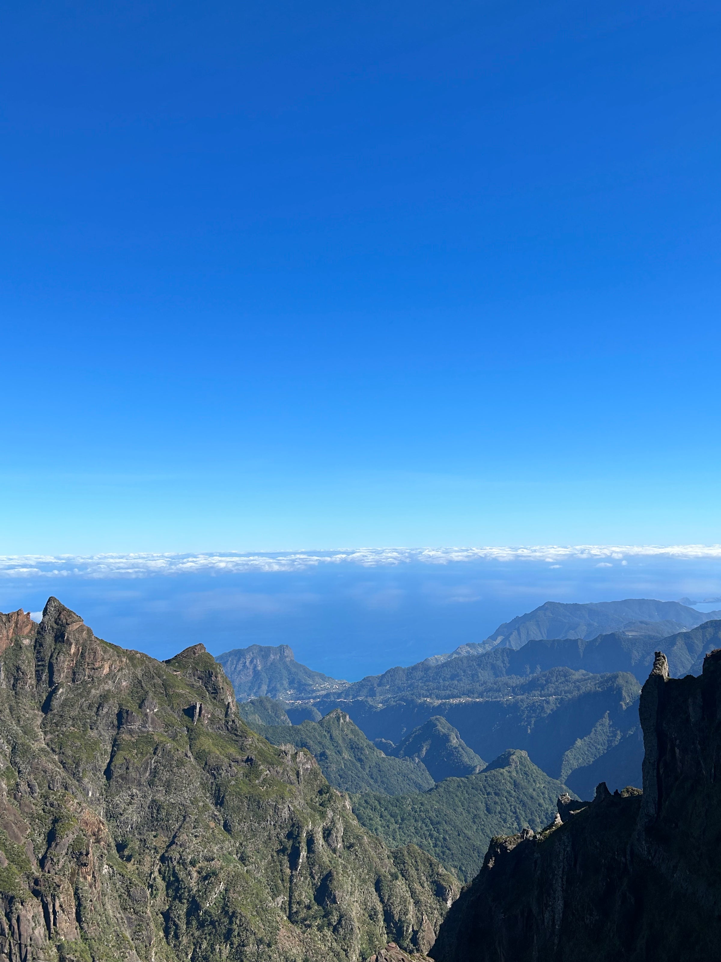 Mountain peaks with green vegetation in the foreground under a clear blue sky, overlooking a distant ocean horizon with a line of clouds above the water.