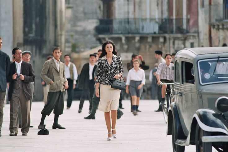 A woman in a polka-dot blouse and cream skirt walks confidently down a city street lined with old buildings, as several men and boys, some on bicycles, watch her. A vintage car is parked nearby.