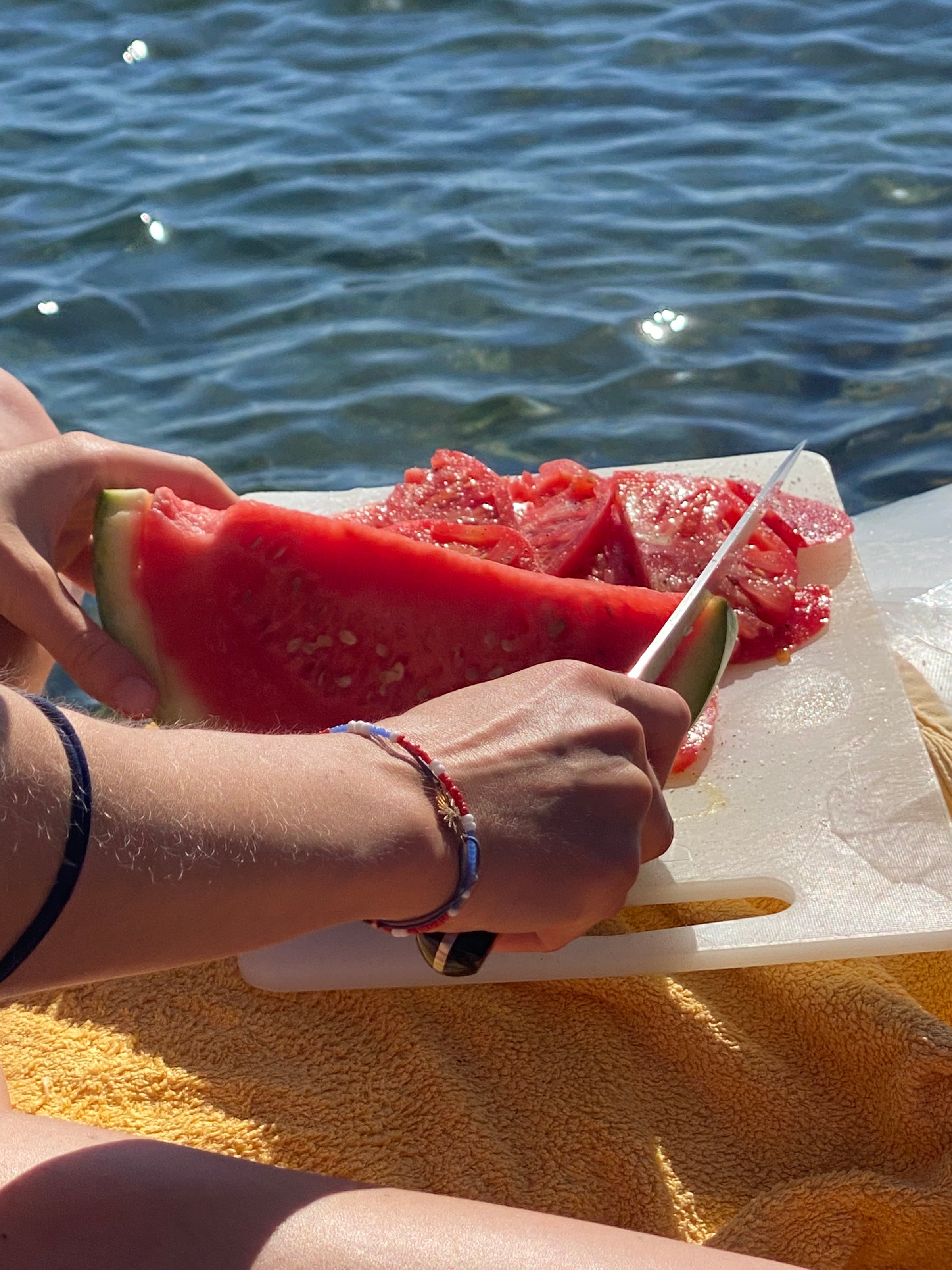 A person slices a large piece of watermelon on a cutting board by the water, with sunlight reflecting on their hands and the fruit. The scene suggests a relaxed outdoor setting, possibly by a lake or the sea.