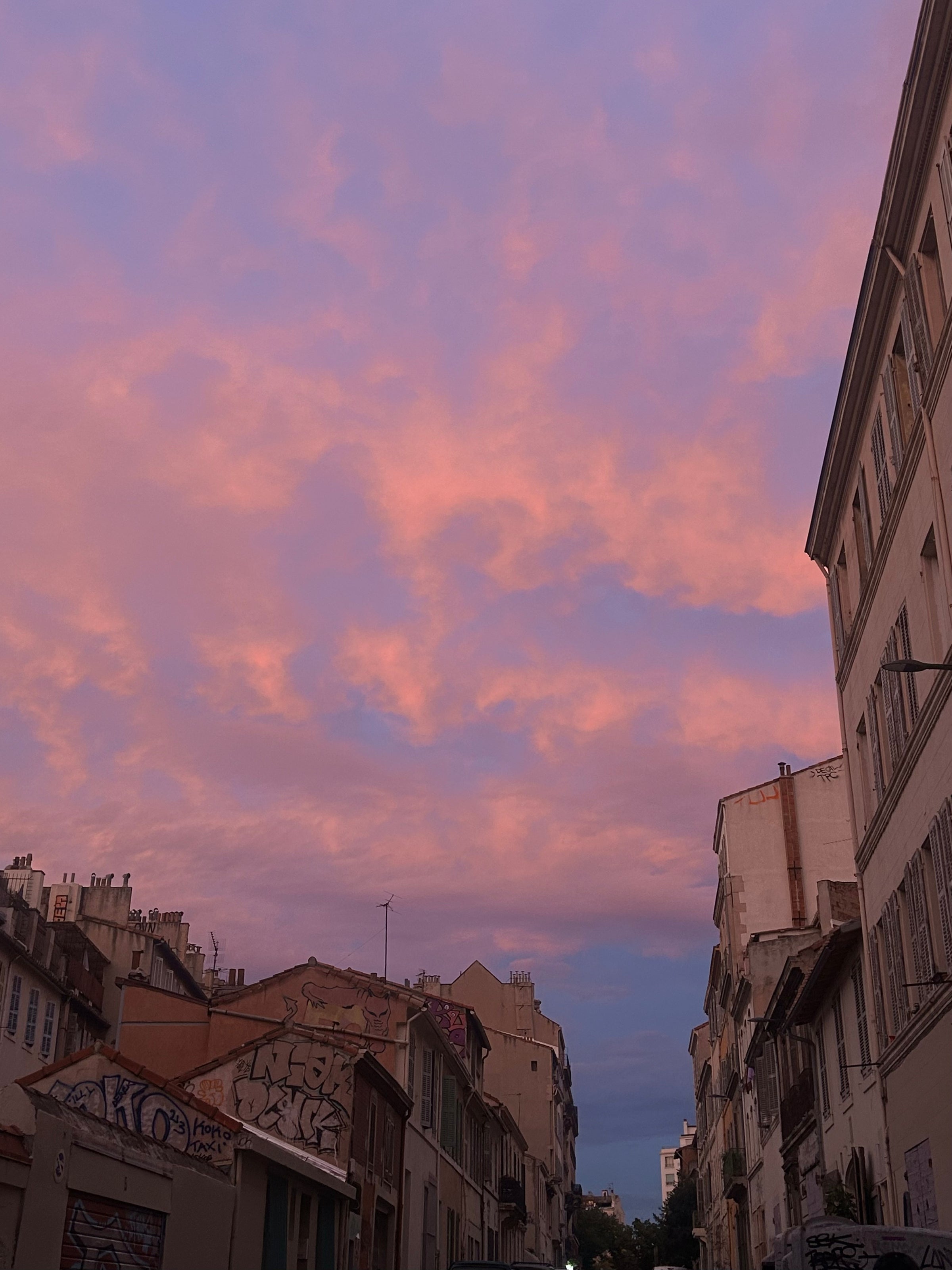 A city street lined with buildings under a vibrant sky at sunset, filled with pink and purple clouds. Some building walls feature graffiti. The scene feels calm and atmospheric.