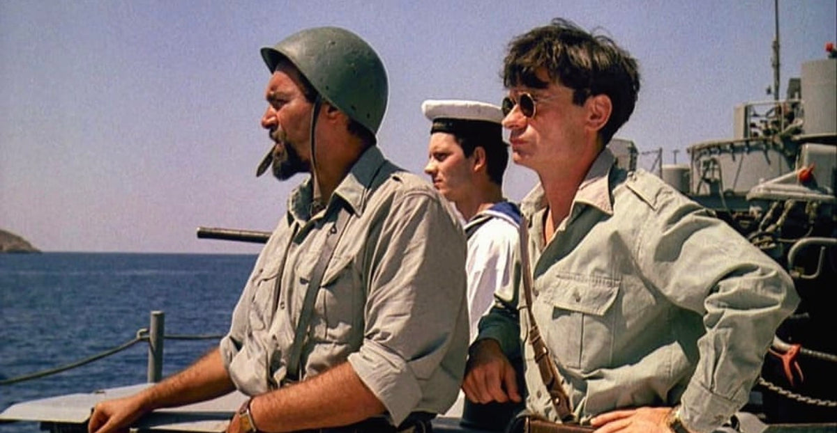 Three men in military-style clothing stand on a ships deck by the sea. One wears a helmet and smokes a cigar, another wears a sailor hat, and the third wears glasses and a green shirt. The sky is clear and sunny.