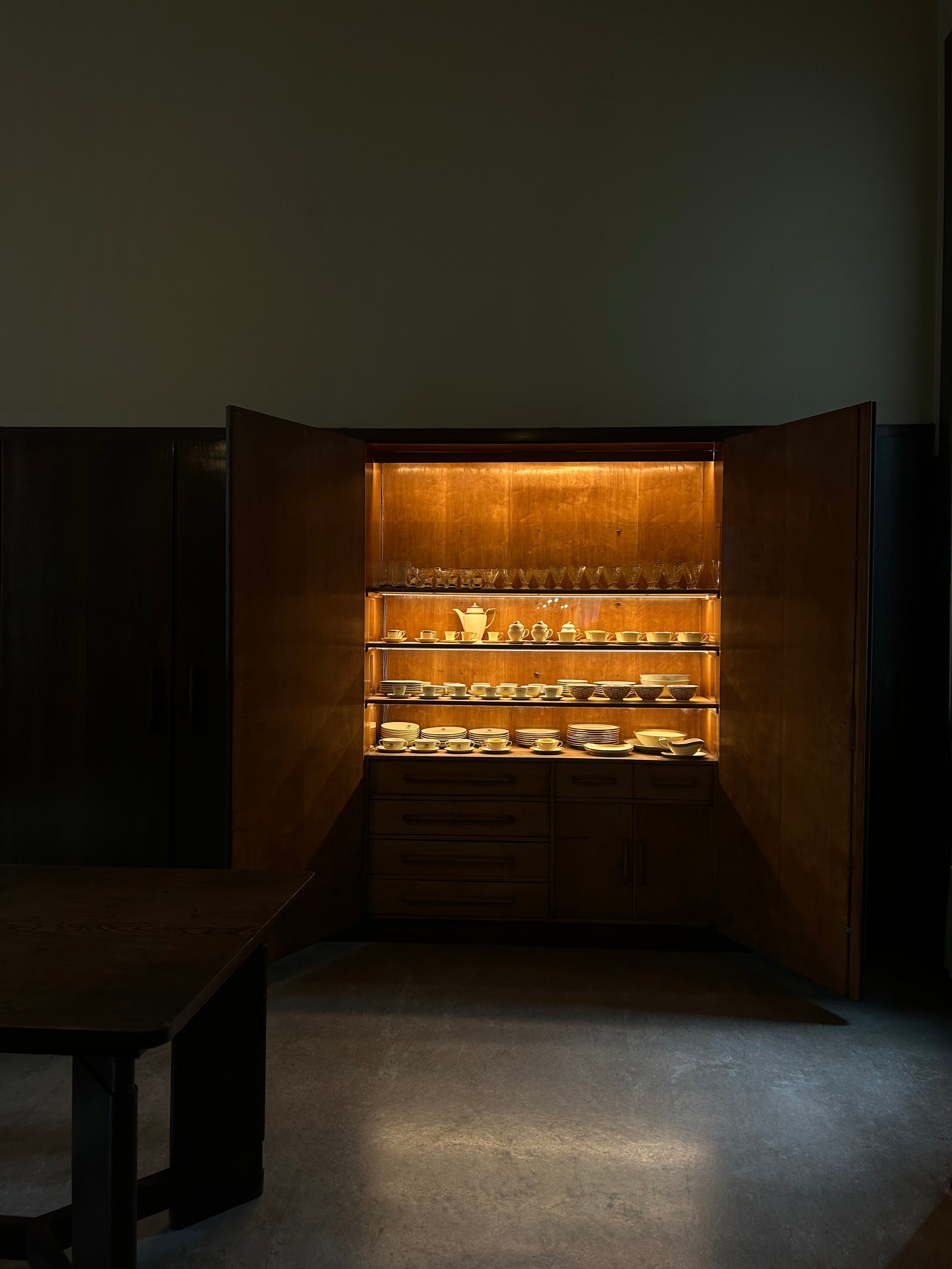 A wooden cabinet with its doors open displays neatly arranged dishes and bowls on illuminated shelves in a dimly lit room.