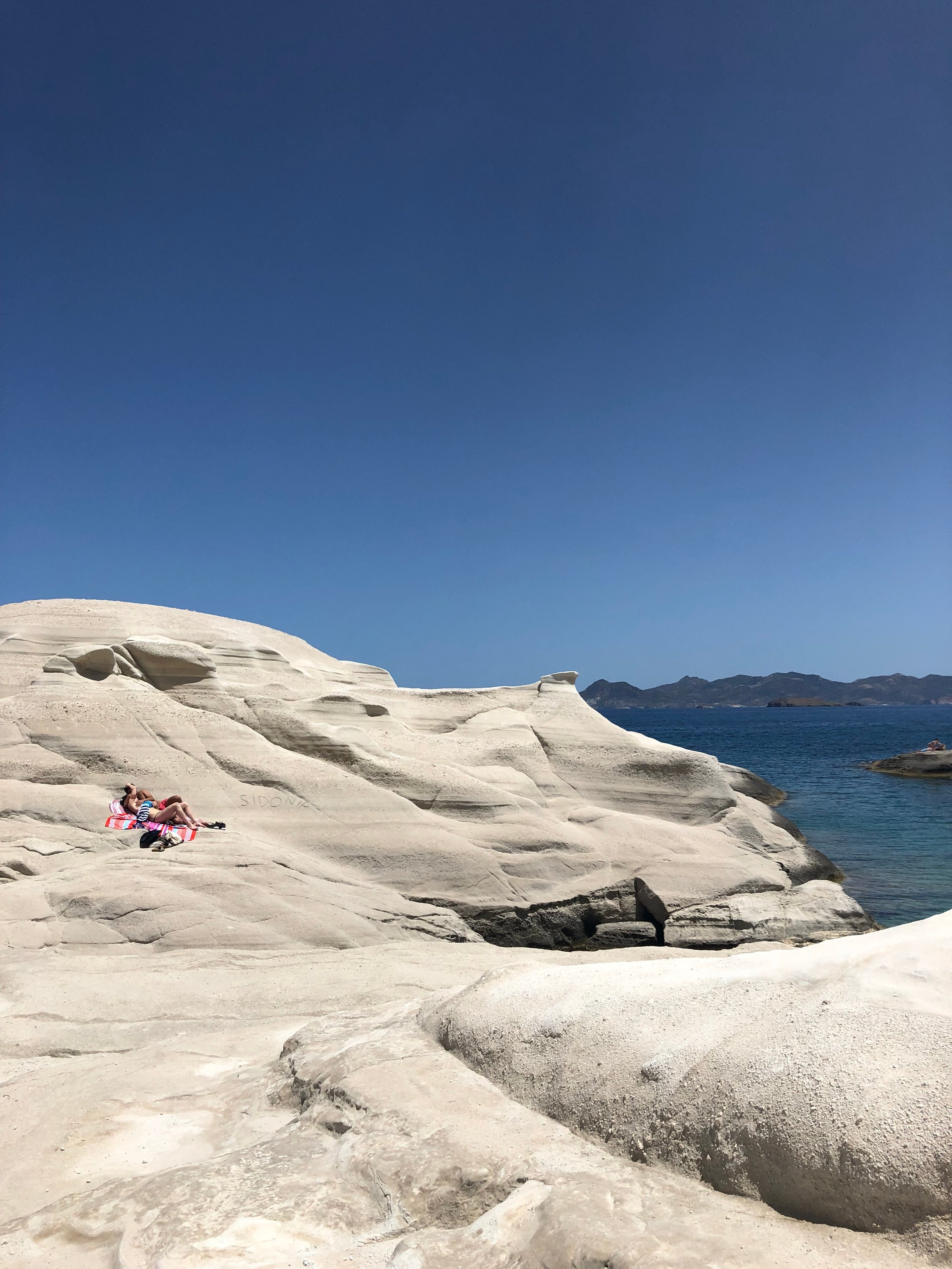 Two people sunbathe on smooth, white rock formations near clear blue water under a bright, cloudless sky, with distant mountains on the horizon.