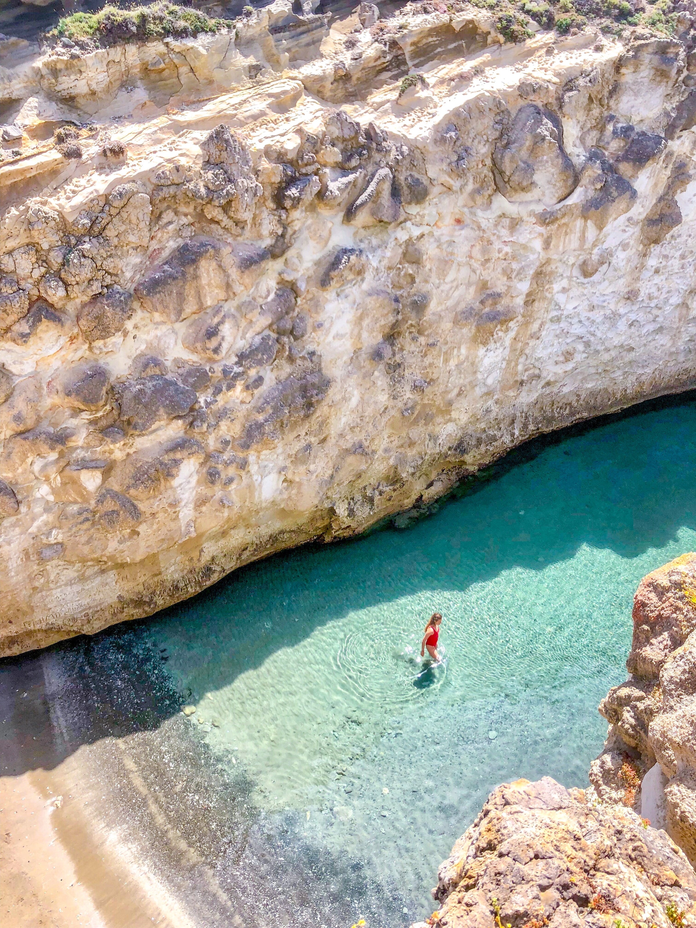 A person wades in clear turquoise water at the base of high, rocky cliffs, with sunlight casting shadows and illuminating the stone and water.