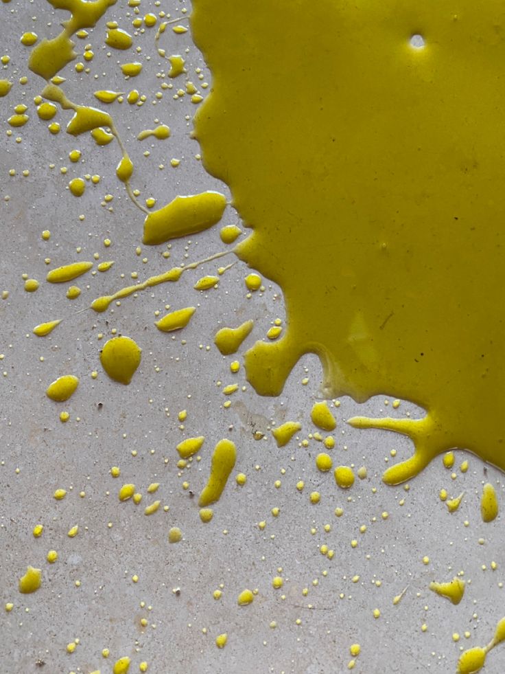 Yellow liquid, possibly oil, is spilled and splattered on a light-colored, textured surface. Droplets and streaks of the liquid create an abstract pattern across the image.