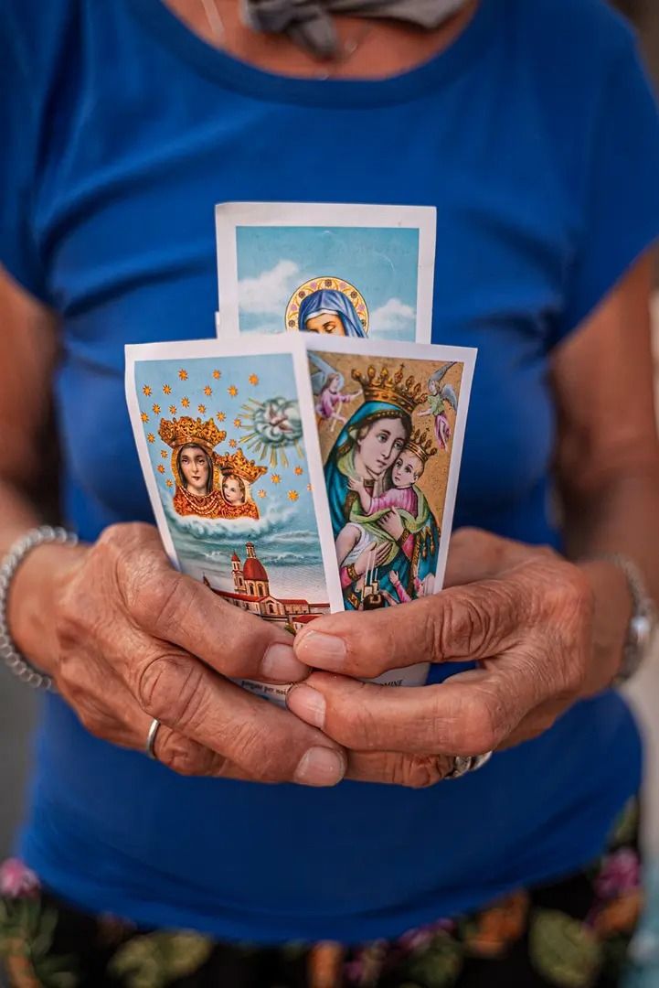 An elderly person with wrinkled hands holds three colorful religious cards, each depicting images of the Virgin Mary, against a background of a blue shirt.