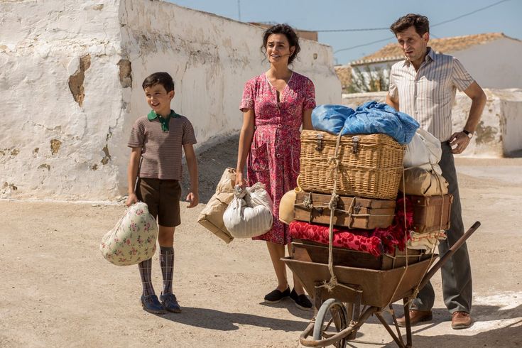 A boy, a woman in a red dress, and a man stand outdoors next to a wheelbarrow loaded with baskets, bags, and blankets. The setting is sunny, with whitewashed buildings in the background.
